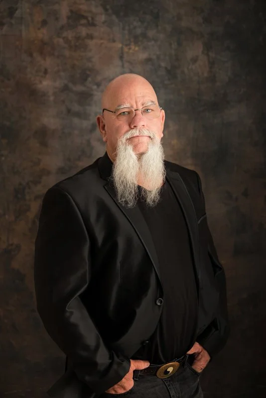 A portrait of a bald man with a long white beard, wearing glasses, a black shirt, and a black blazer, standing against a dark textured background.