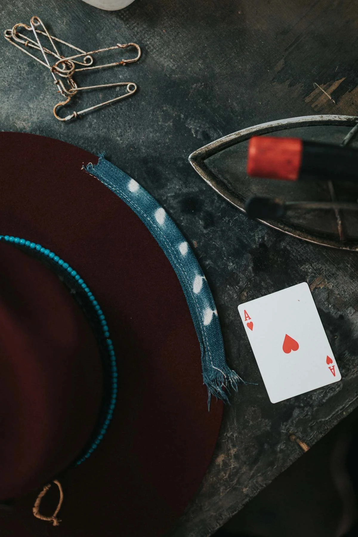 Close-up of a black table with small rusted safety pins, a red hat with a teal beaded band, a red and white bracelet, an iron, and a playing card indicating the Ace of Hearts.
