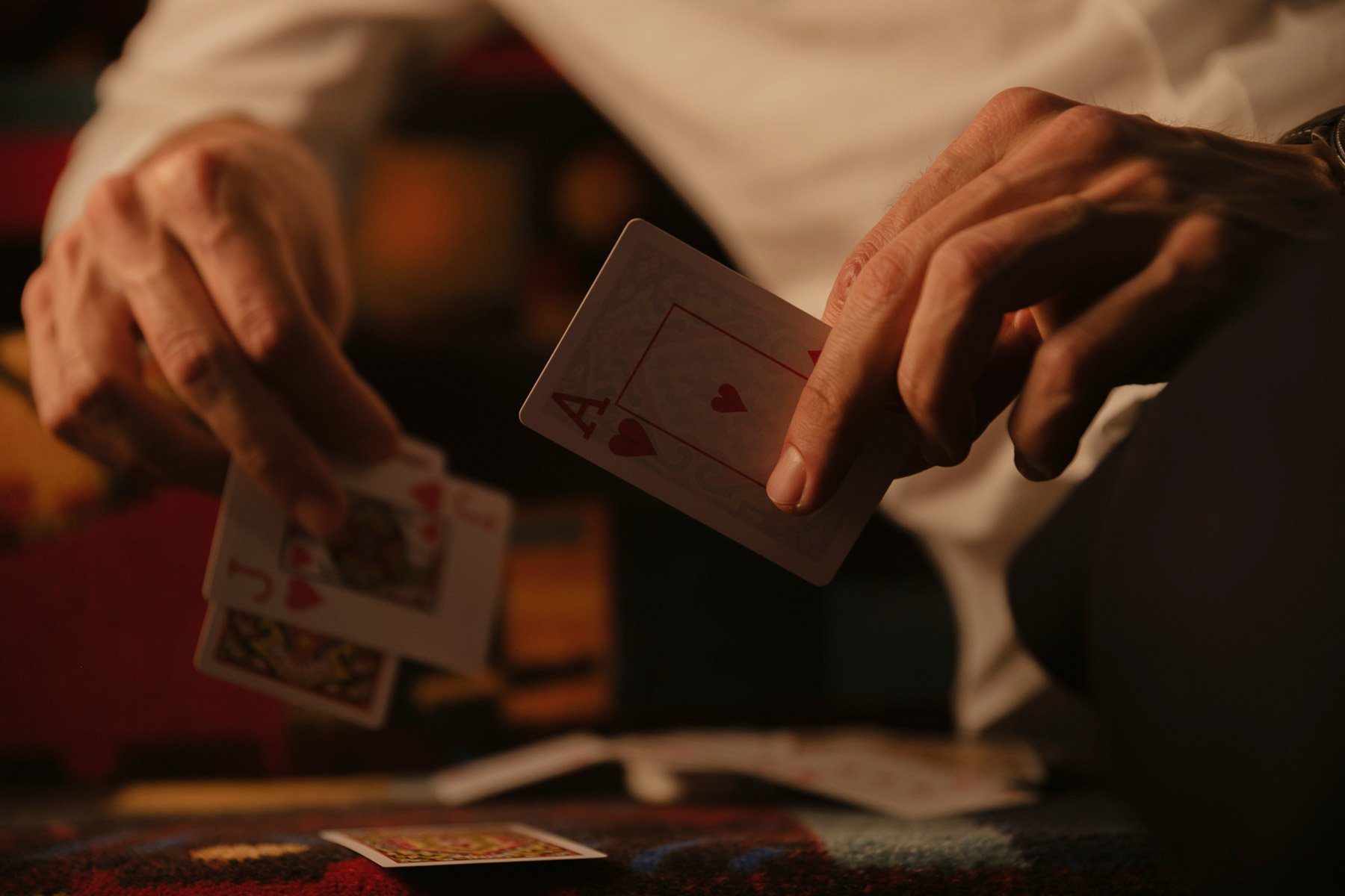 Close-up of a hand holding two playing cards, the Ace of Hearts and the Jack of Hearts, with additional cards visible on a table in the background.