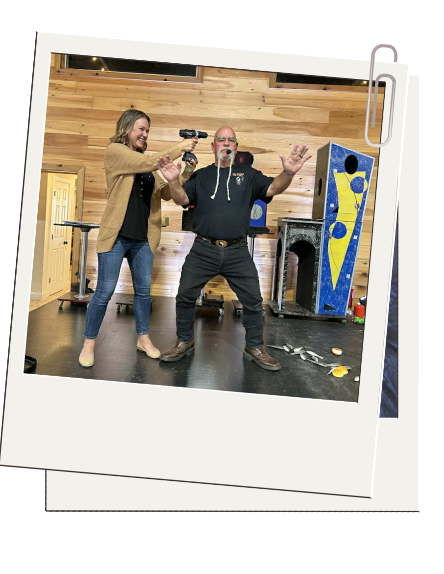 A woman holding a power drill and a man with a beard and glasses, wearing a headset and a black shirt, participating in an activity on a stage with wooden paneling, surrounded by colorful play sculptures, debris, and a fire extinguisher.