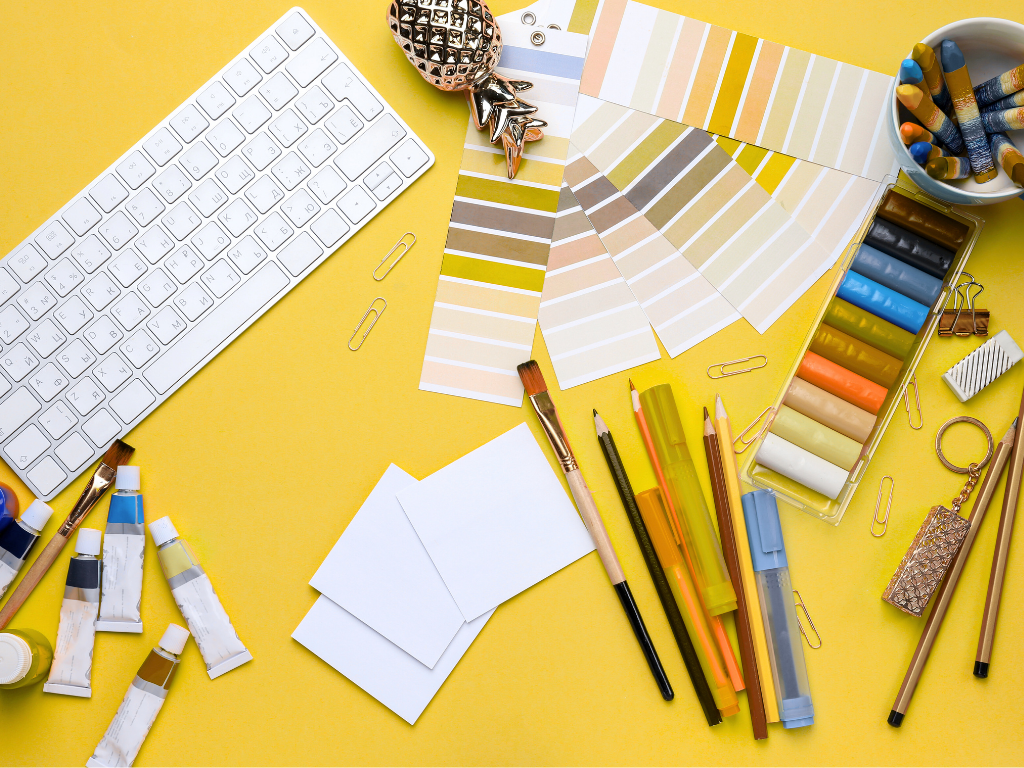 Workspace with white keyboard, paint tubes, colored pencils, paint brushes, colorful pastels, color swatches, paper clips, and stationery against a yellow background.