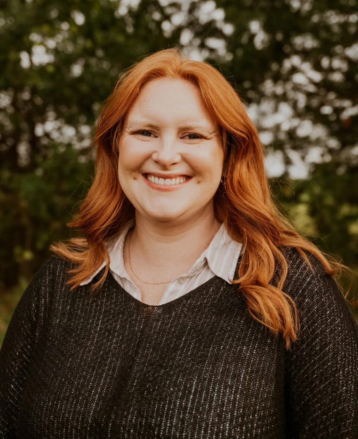 A woman with red hair, smiling, wearing a black sweater over a white collared shirt, outdoors with trees in the background.