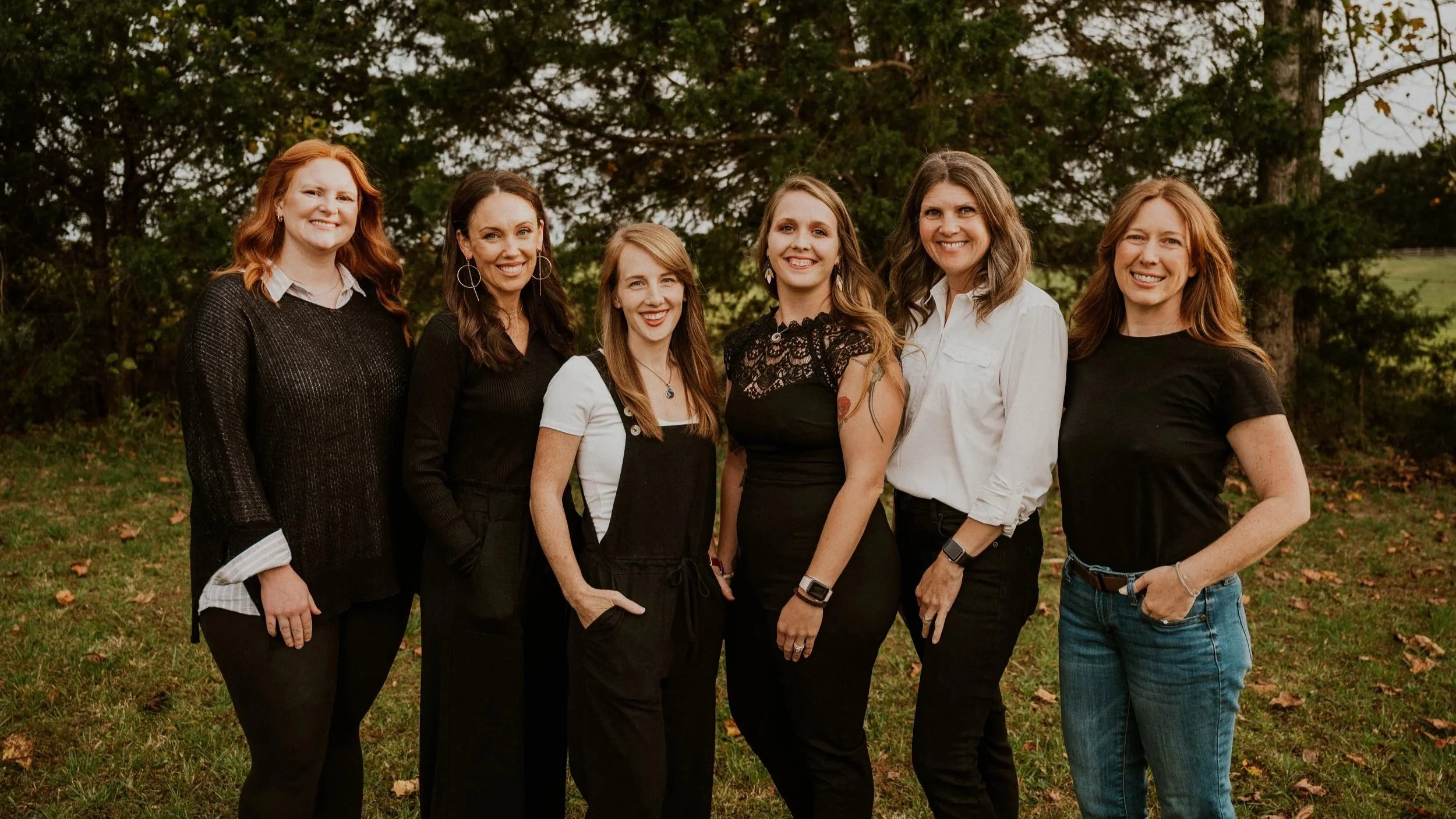 Six women standing outdoors on grass in front of trees, smiling at the camera.