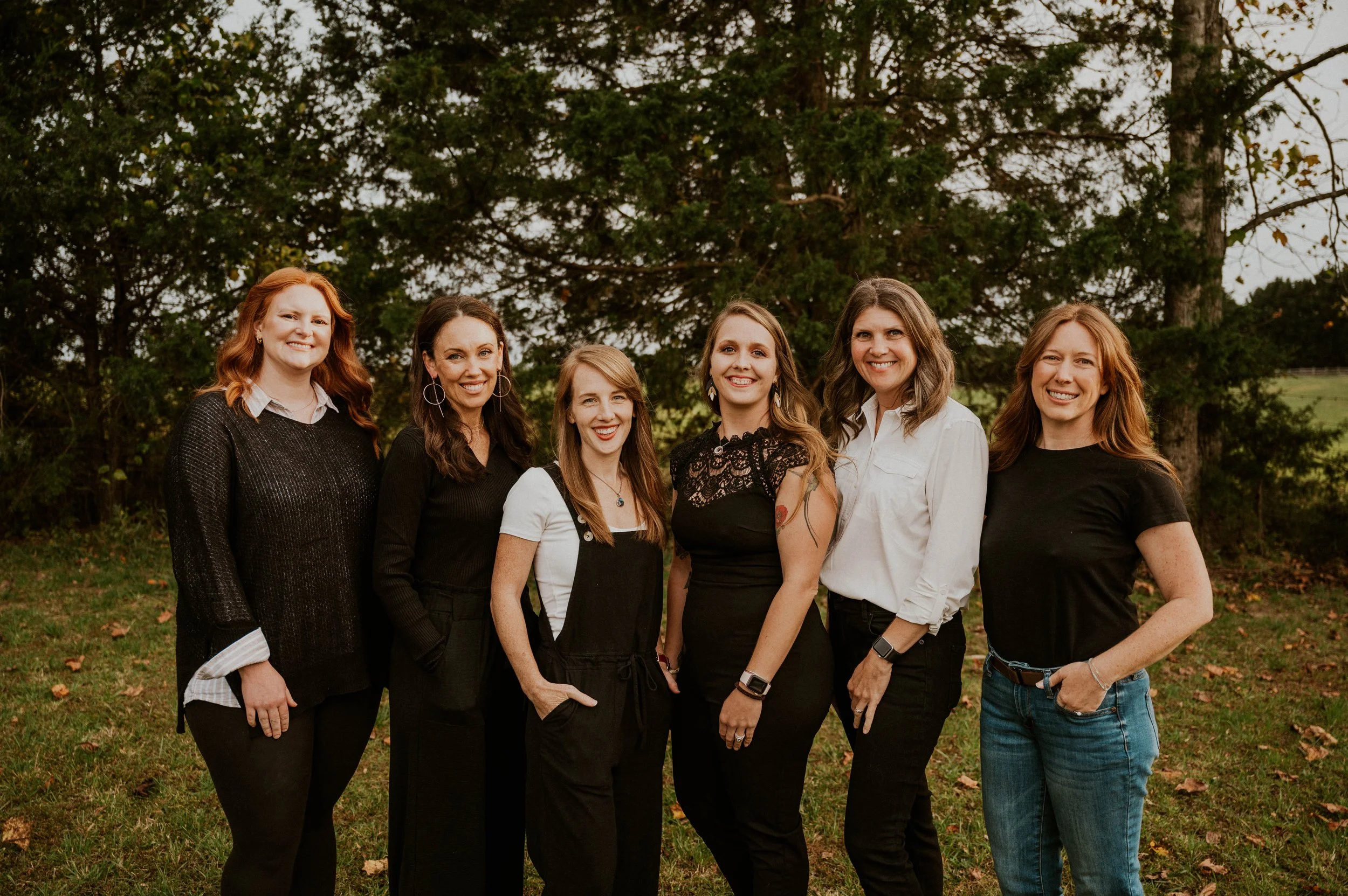 Group of six women standing outdoors in front of trees, smiling at the camera.
