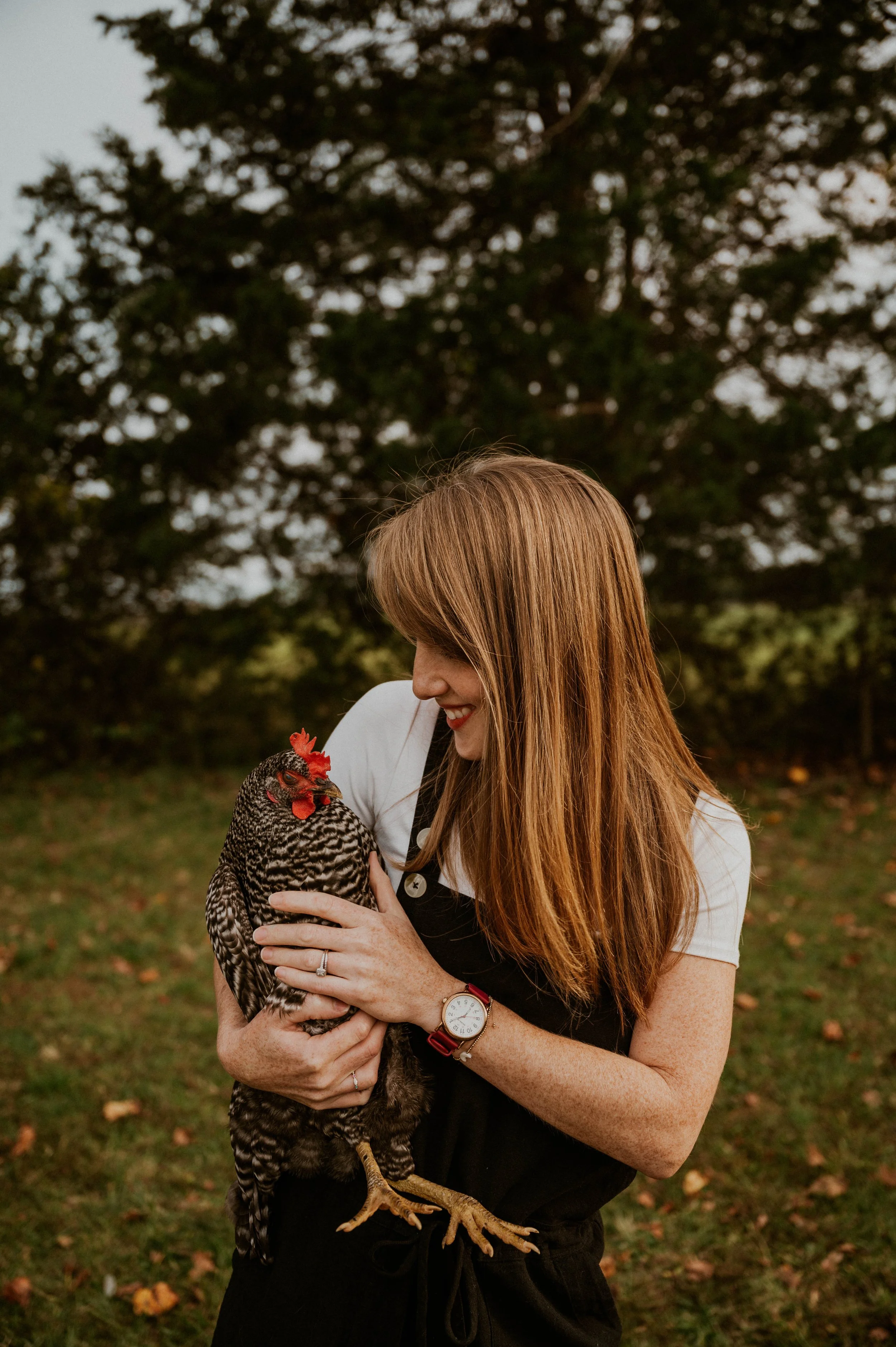 A woman with long red hair smiling and holding a hen outdoors on a grassy area with trees in the background.
