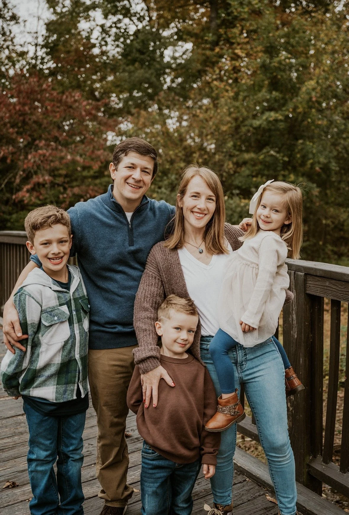 A family of five on a wooden bridge outdoors during autumn. The parents are smiling with three children, a girl and two boys, all dressed in casual autumn clothing. The background features trees with fall foliage.