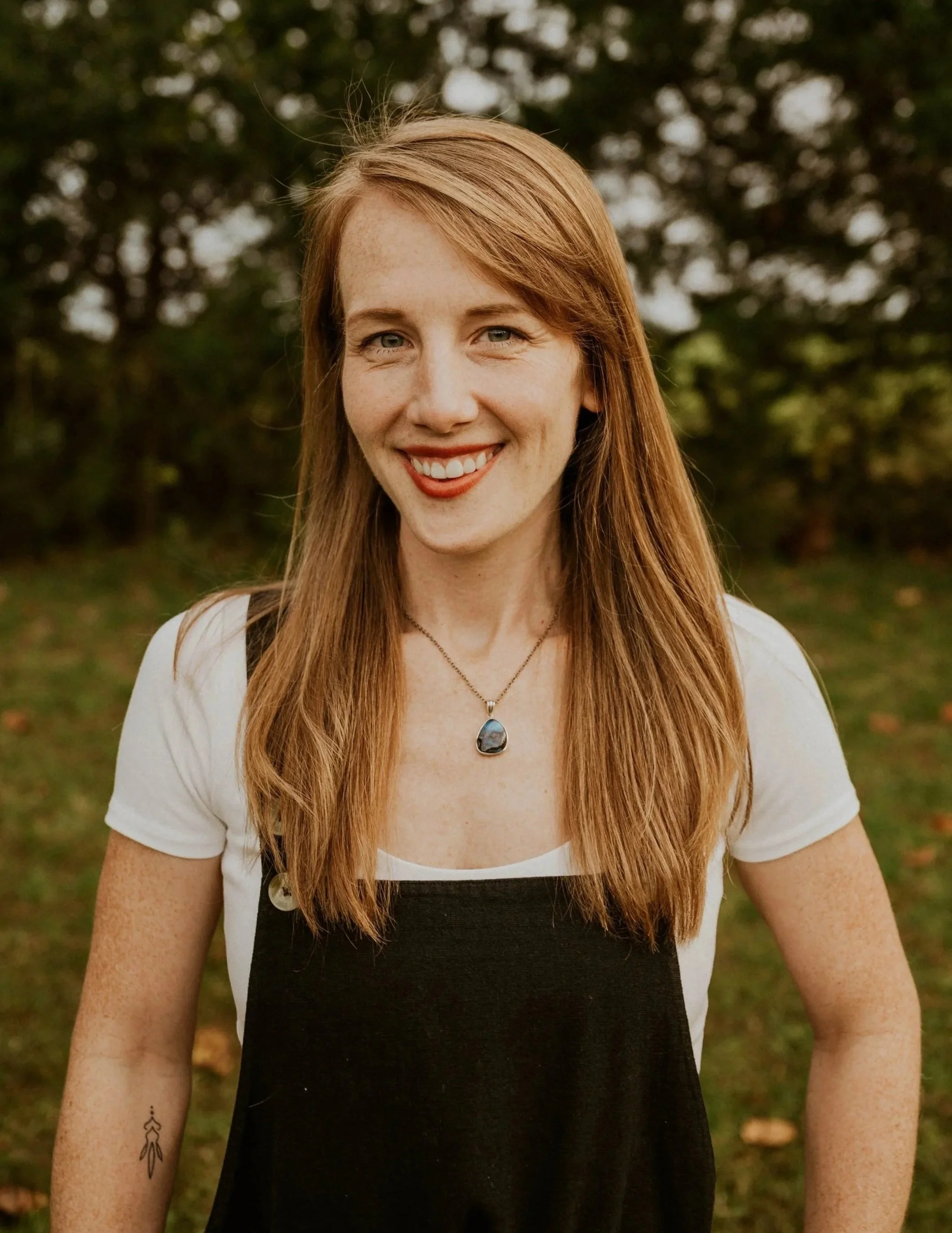 A smiling woman with long red hair, wearing a white t-shirt with black dress over it, a necklace with a blue pendant, and standing outdoors in a park with green trees in the background.