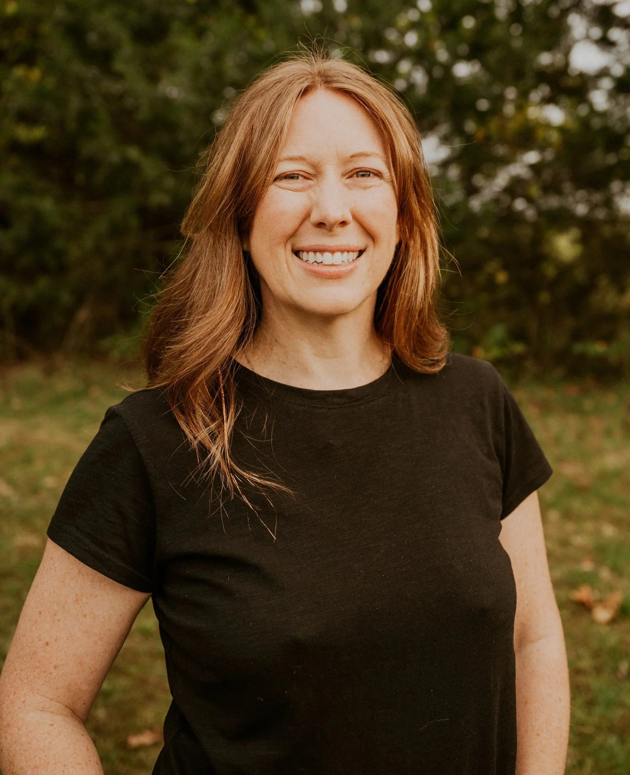 A woman with red hair outdoors in front of trees, smiling and wearing a black t-shirt.