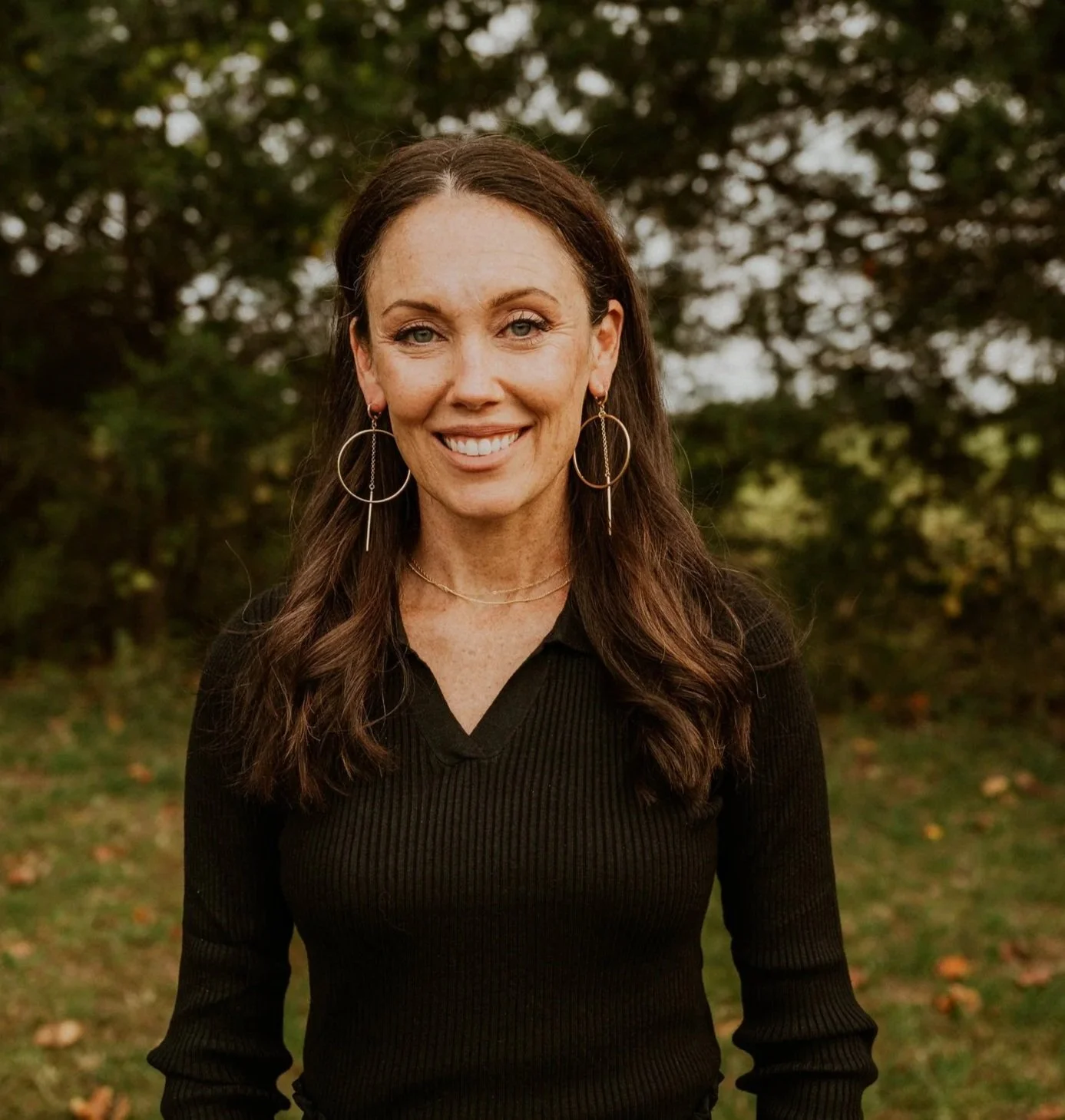 A woman with long brown hair, wearing a black sweater, large hoop earrings, and a delicate necklace, smiling outdoors with trees in the background.