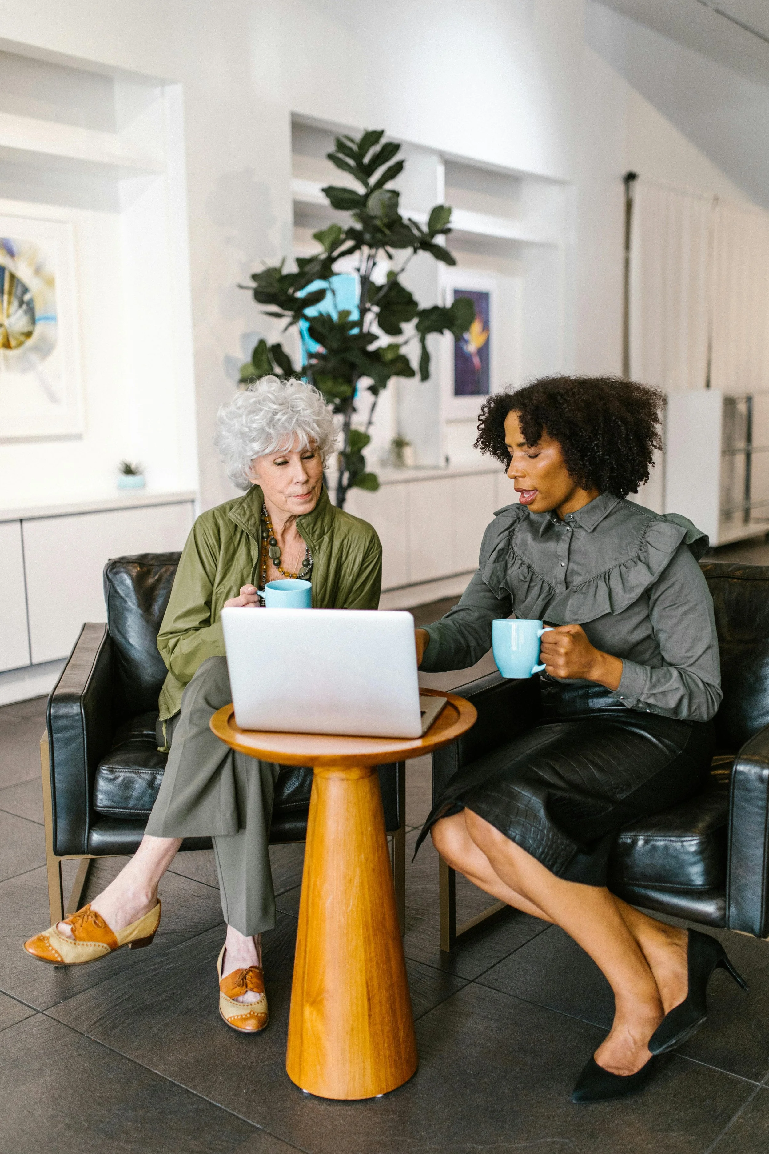 Two women sitting in a modern office environment, engaged in conversation while looking at a laptop screen. They are holding coffee mugs and seated on black armchairs with a small wooden table between them. There is art on the wall and a plant in the background.