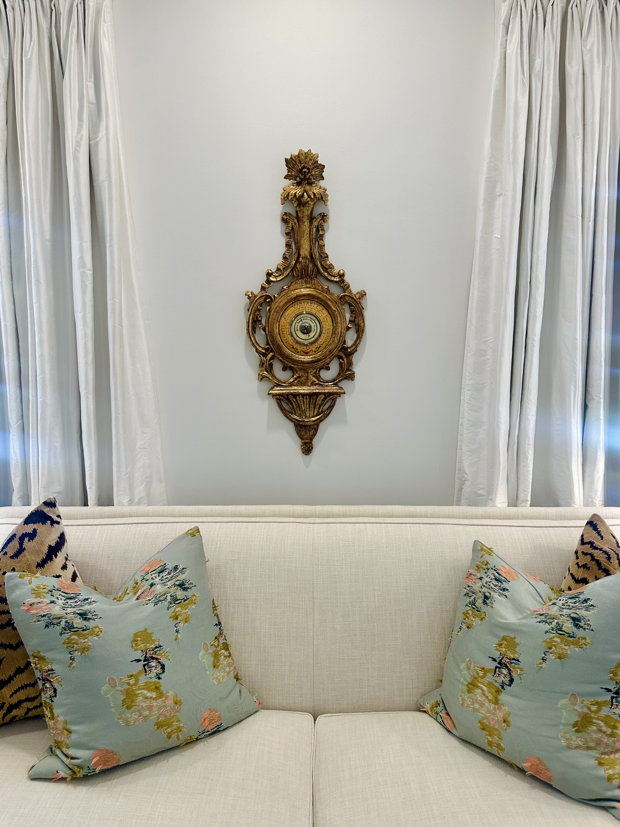 Elegant living room with a white sofa, floral and patterned pillows, white curtains, and an ornate gold wall decoration.