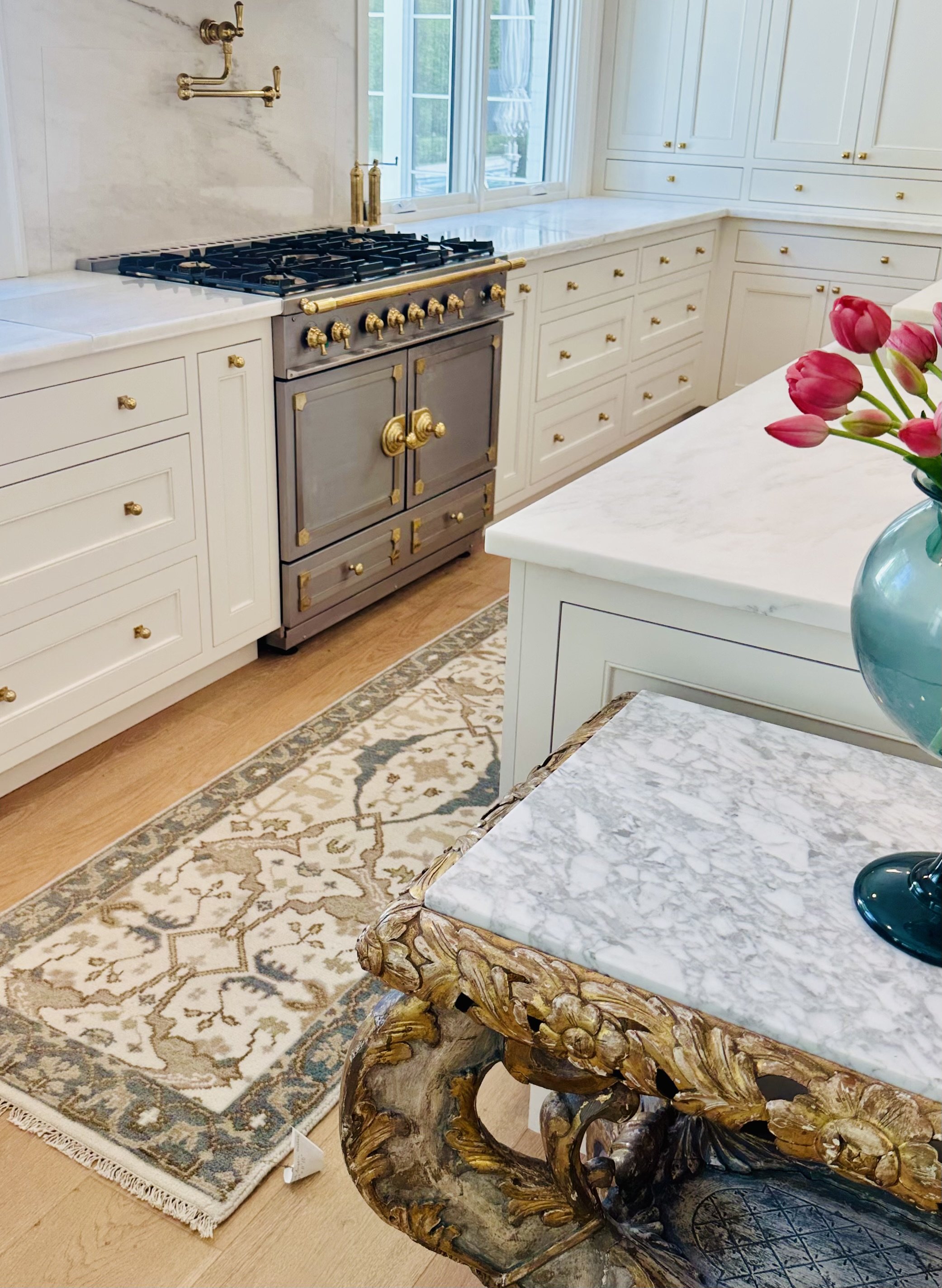 Elegant kitchen with a vintage-style stove featuring gold accents, white cabinetry, marble countertops, a decorative area rug, and a vase of tulips on a marble-topped table with ornate carvings.