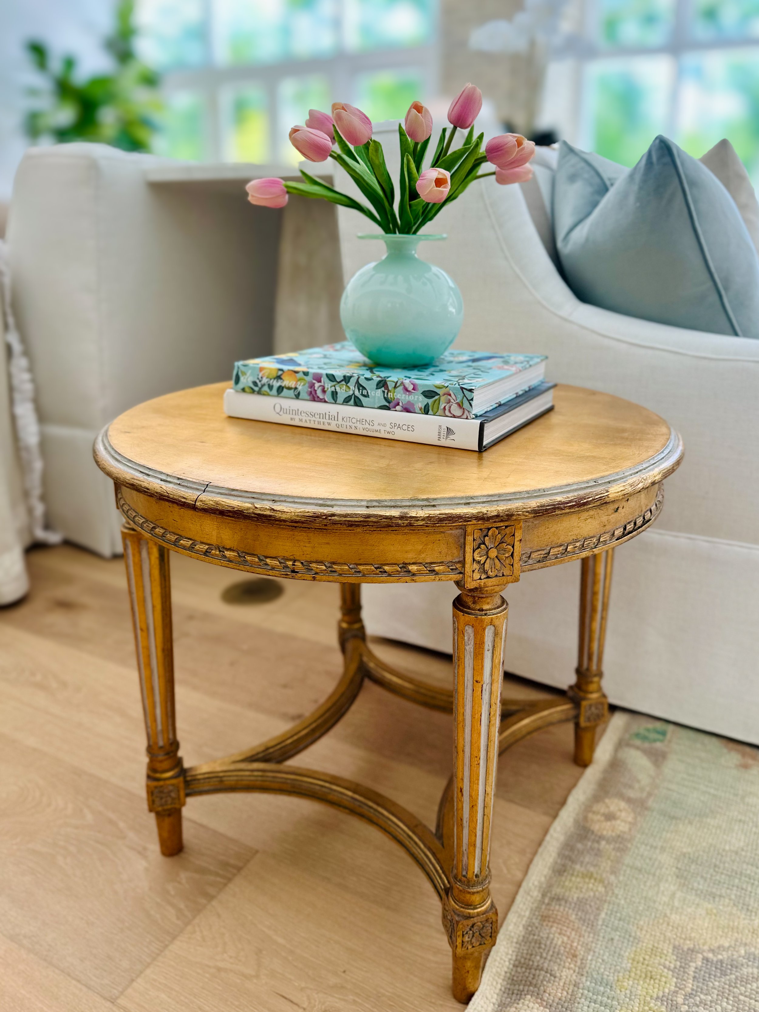 Round wooden side table with carved floral details, featuring a blue vase holding pink tulips and two stacked books, beside a white sofa with blue pillows in a bright living room.