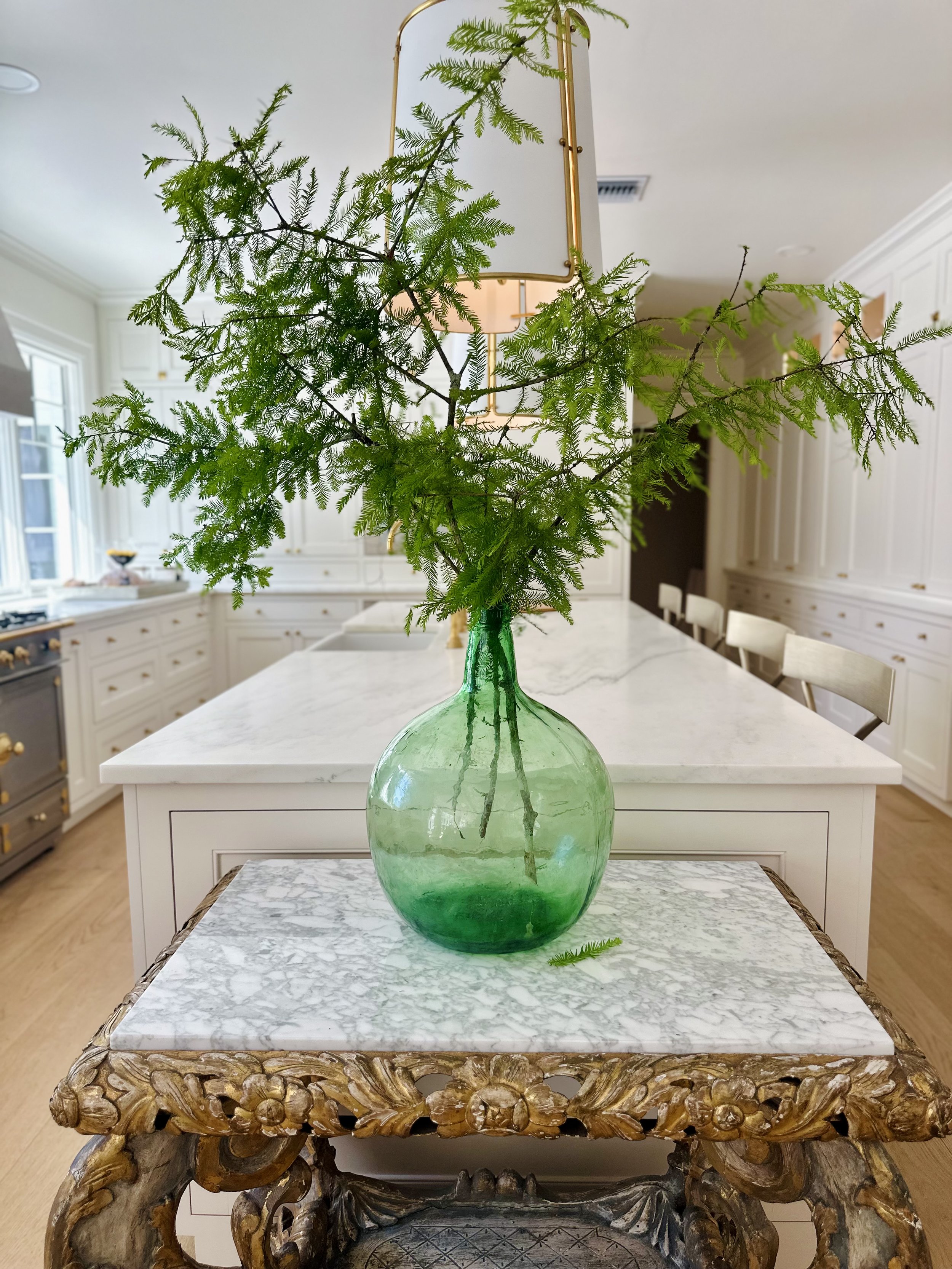 Elegant kitchen with a green glass vase holding green branches on a marble-topped ornate table in front of a kitchen island with white cabinets.