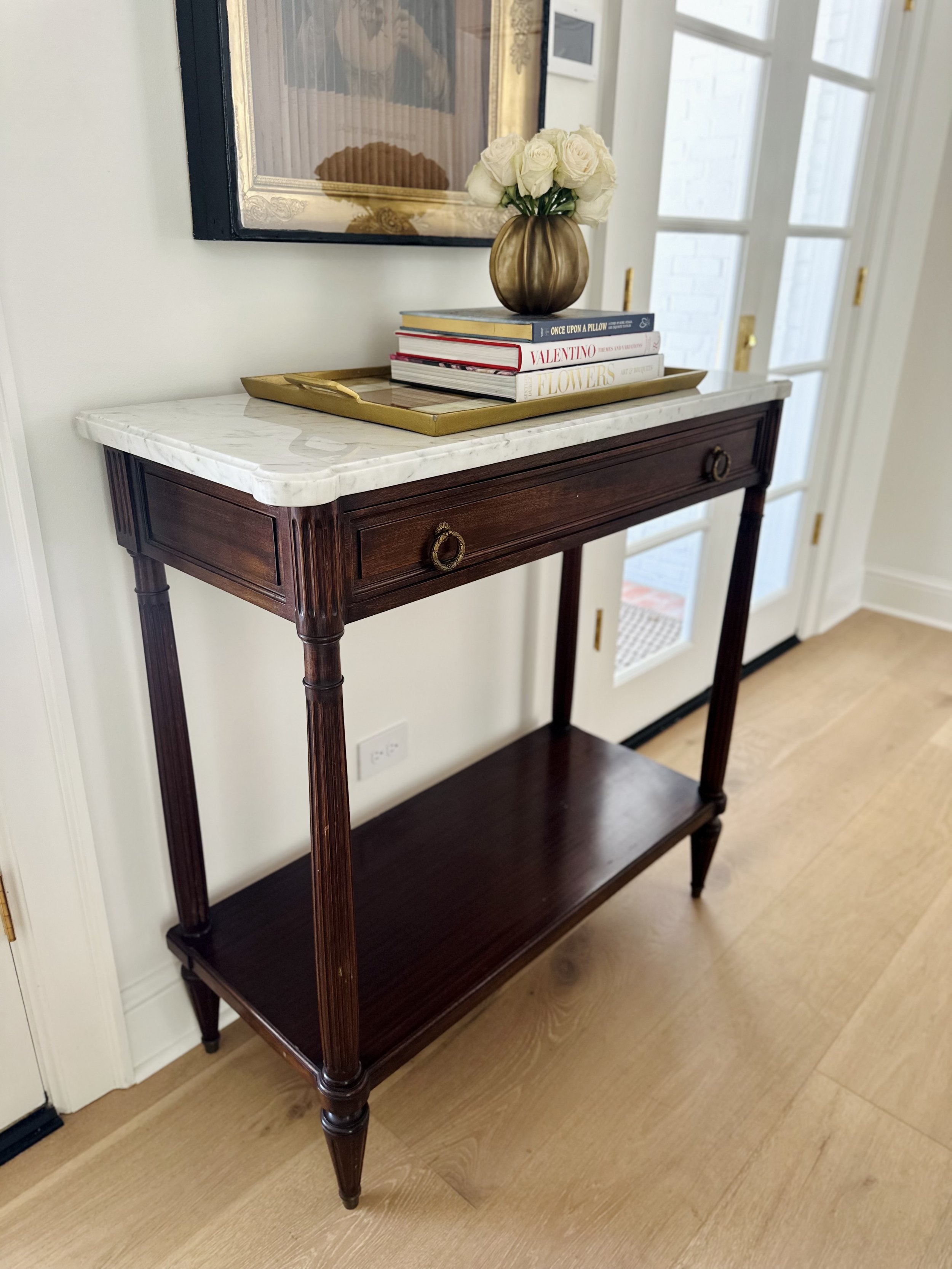A wooden console table with a marble top, features books, a gold-colored vase with white roses, and a decorative tray. A framed picture hangs above the table, next to a white door with glass panels.
