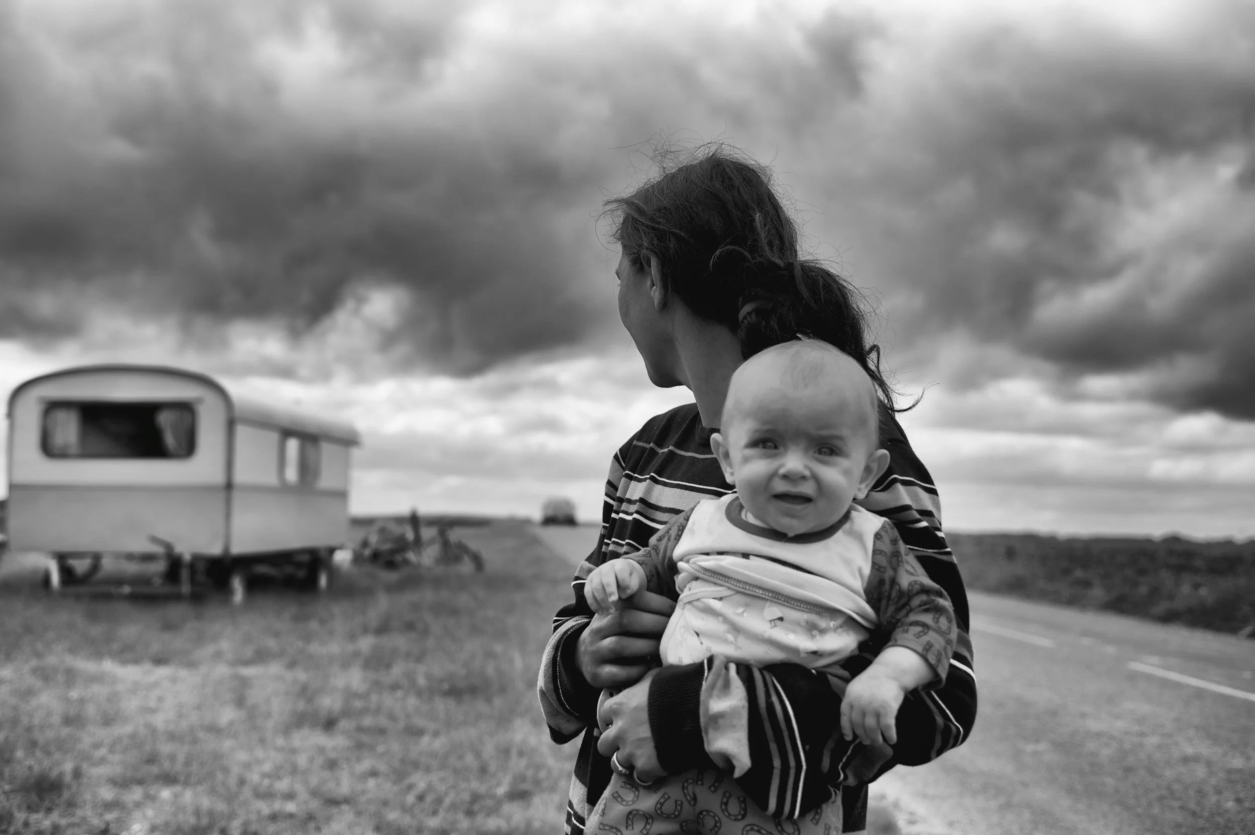 Black and white photo of a woman holding a baby, standing near a road with a caravan in the background under a cloudy sky.