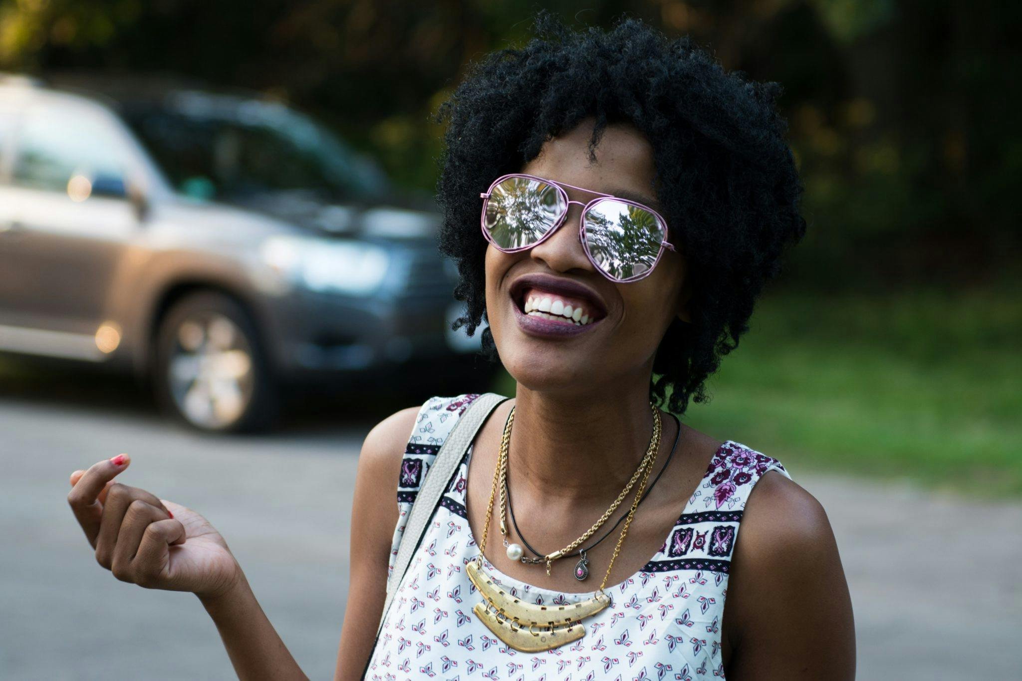 Smiling woman wearing reflective sunglasses, patterned dress, and layered necklaces outdoors.
