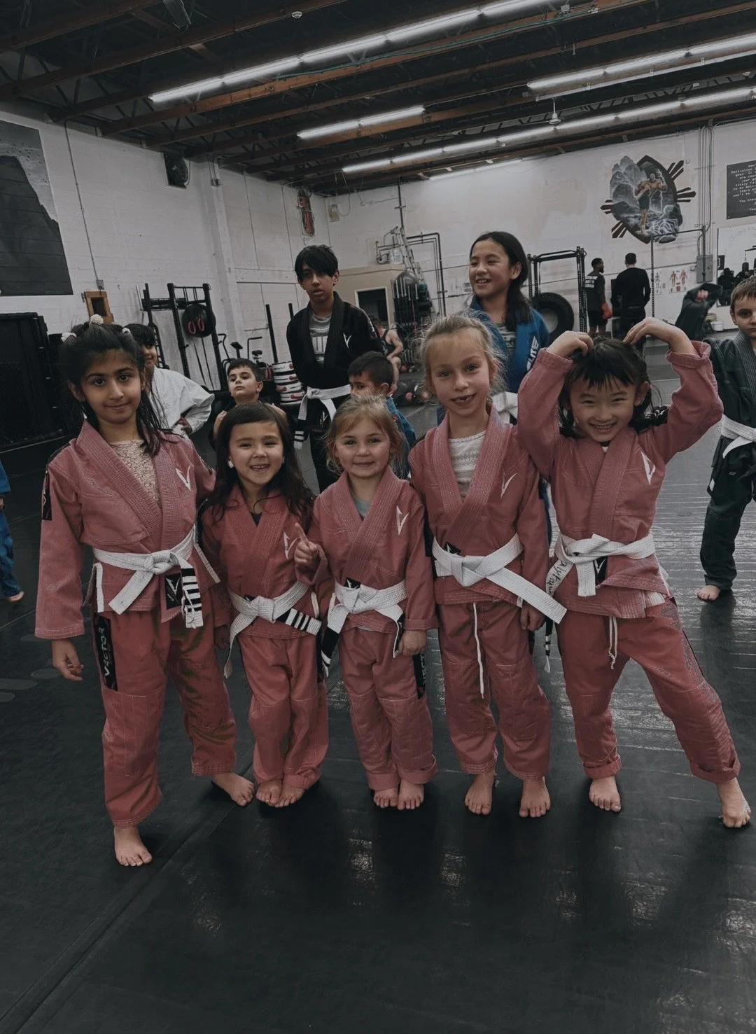 Children in pink martial arts uniforms with white belts posing in a gym, with others in the background.