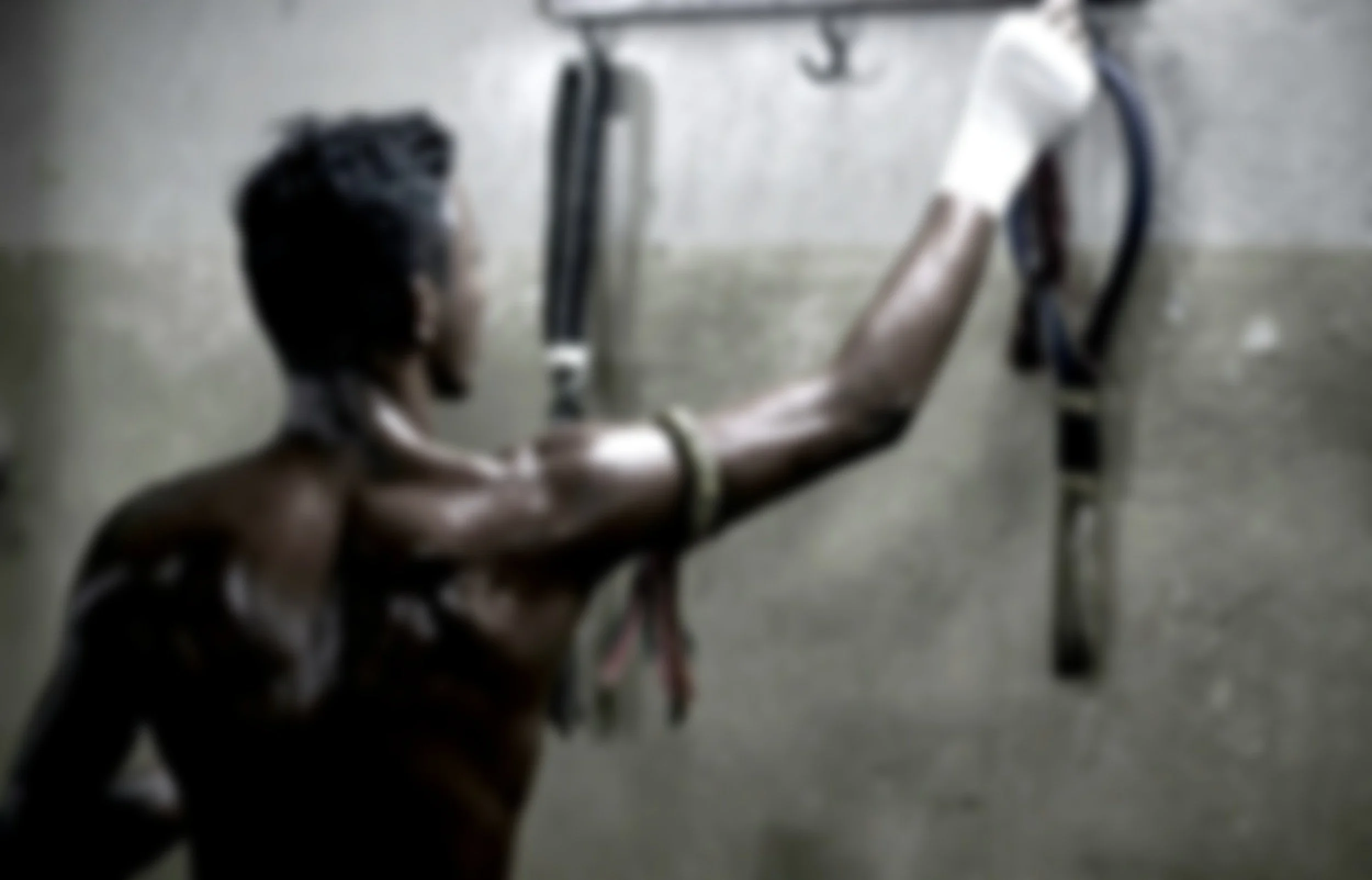 A shirtless man with a beard and a wristband reaches out to grab a pair of metallic crutches mounted on a gray plastered wall.