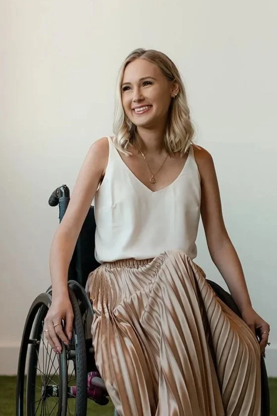 A young woman with blonde hair smiling while sitting in a wheelchair against a plain white wall, wearing a sleeveless cream top and a shiny, pleated beige skirt.