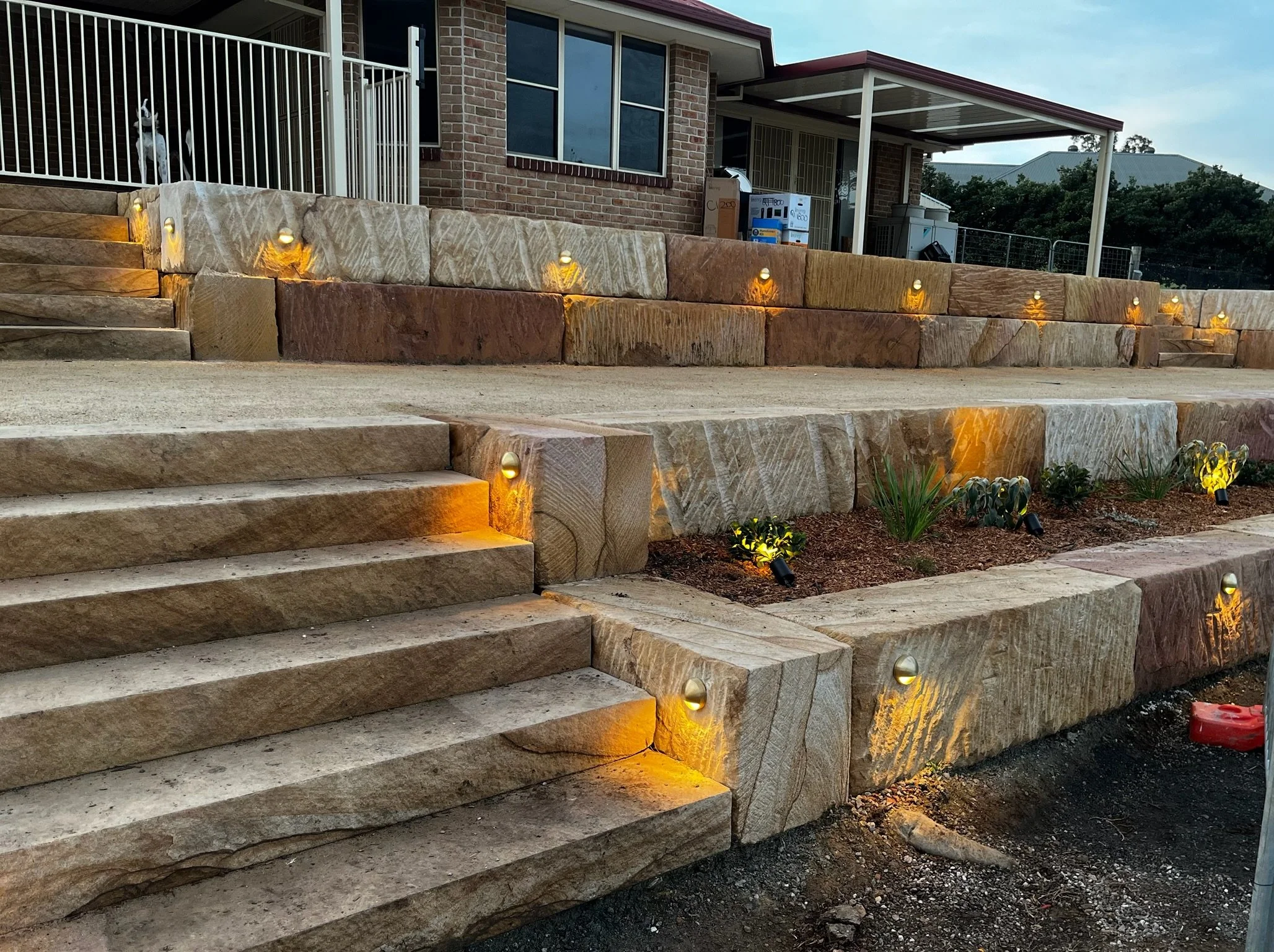 Stone steps with built-in pathway lights, leading to a brick house with large windows and a porch, landscaping with small plants and mulch.