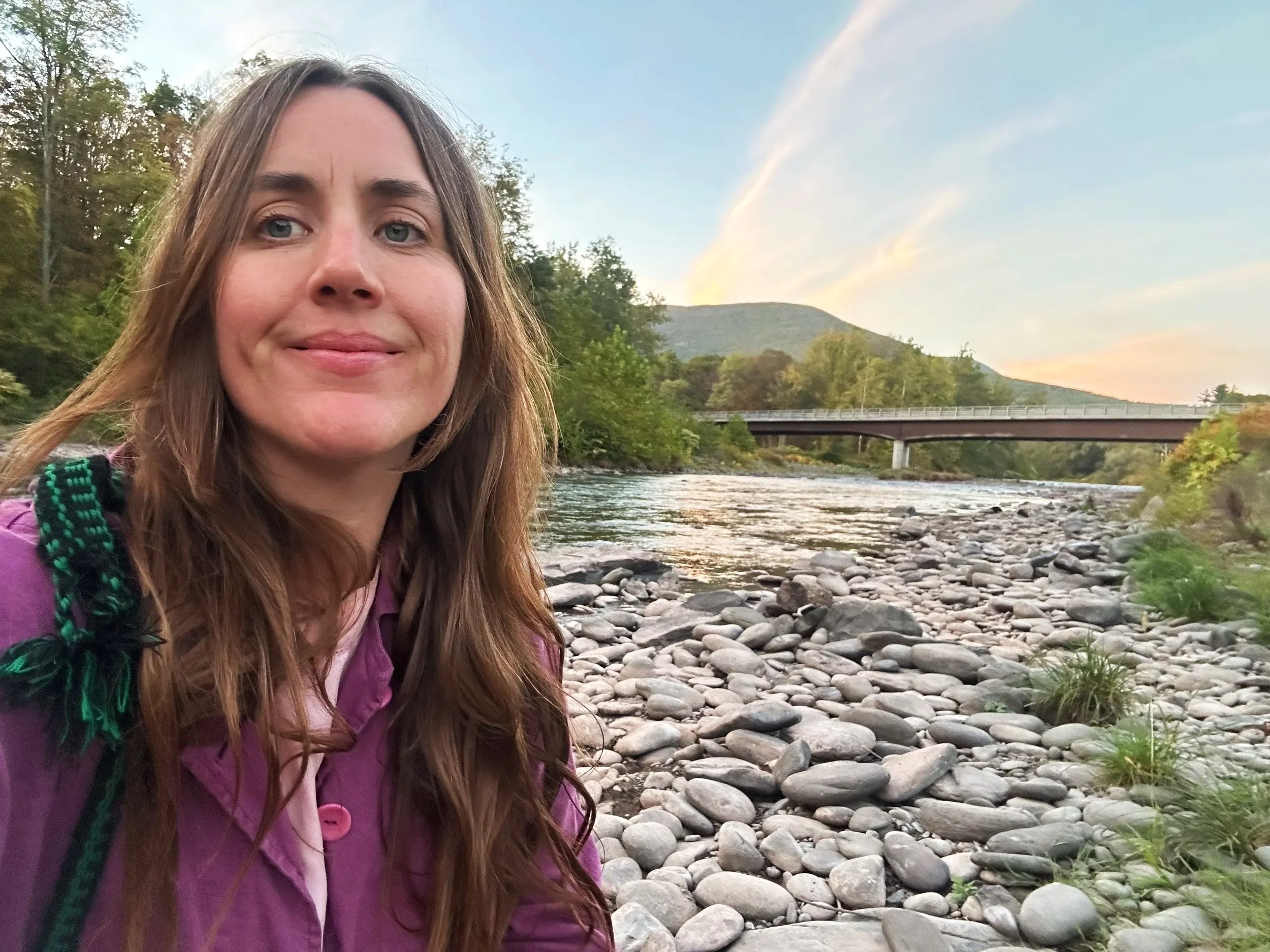 A woman with long light brown hair, wearing a purple jacket and a backpack, standing by a rocky riverbank with a bridge in the background, trees, and mountains during sunset or sunrise.