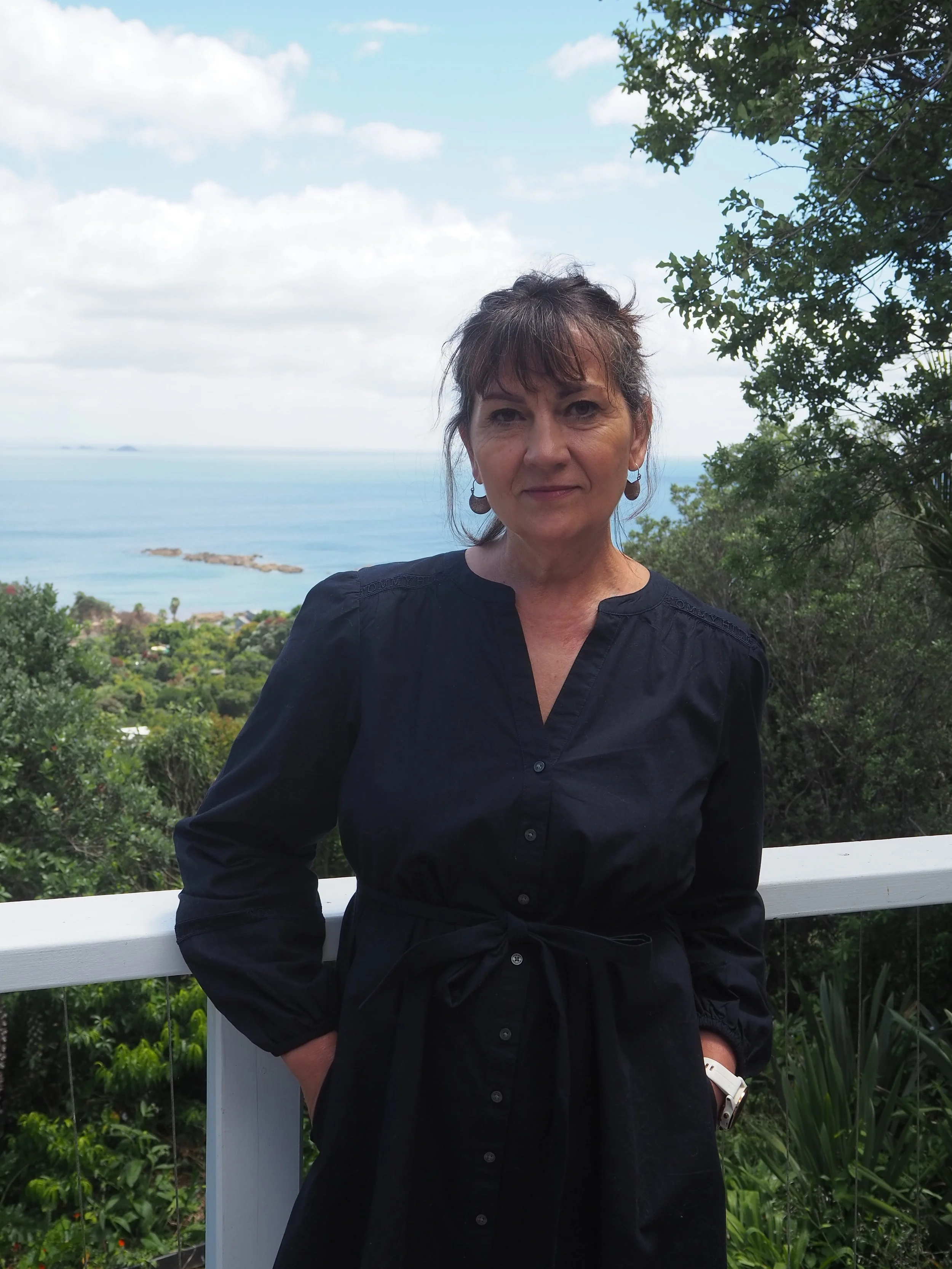 A woman wearing a black shirt standing outdoors on a balcony with a scenic view of the ocean, greenery, and a partly cloudy sky in the background.