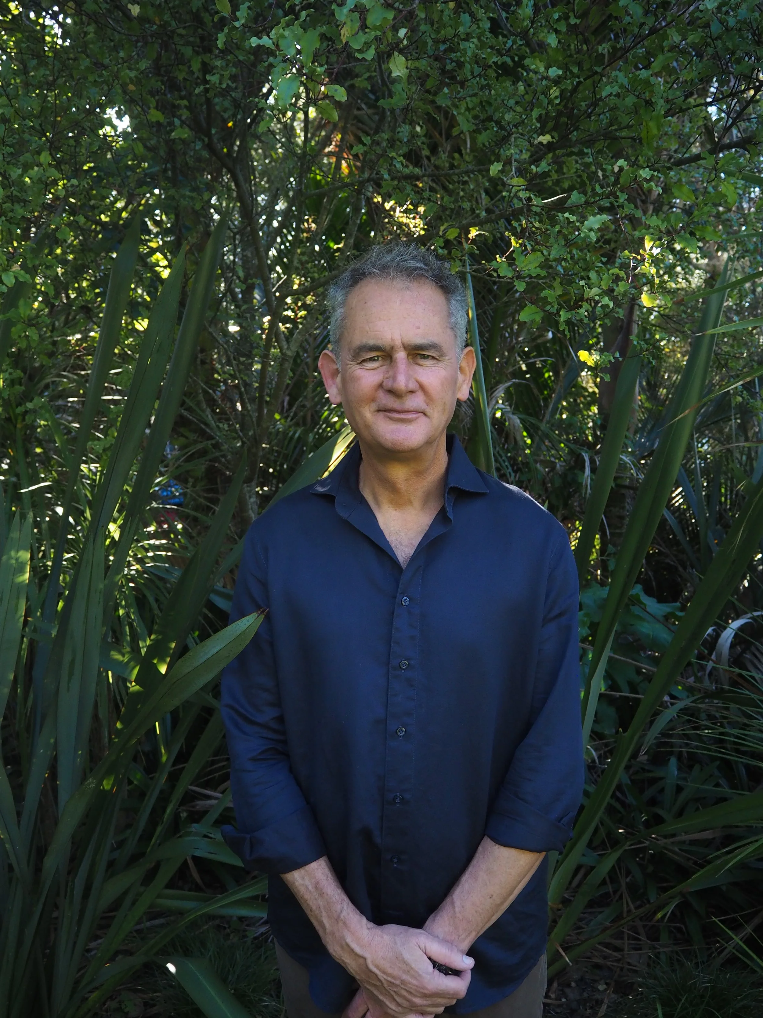 A middle-aged man with short gray hair and wearing a dark blue buttonup shirt standing outdoors in front of dense green foliage.
