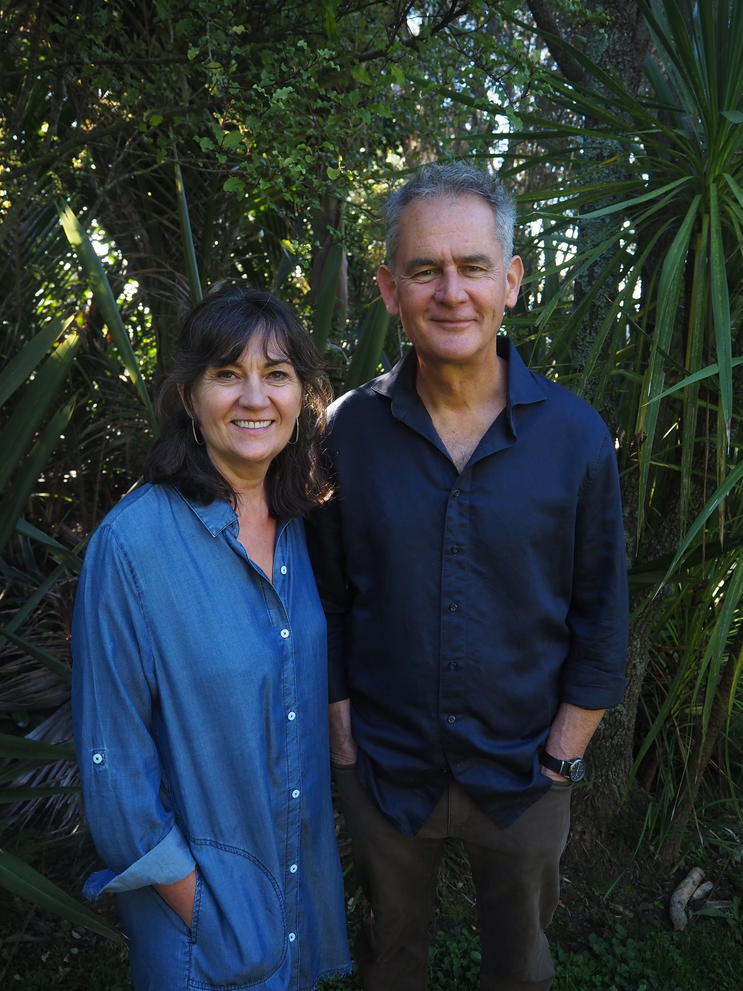 A woman and a man standing outdoors in front of green foliage, smiling at the camera. The woman is wearing a blue denim shirt, and the man is wearing a dark blue shirt with sleeves rolled up and a watch on his left wrist.