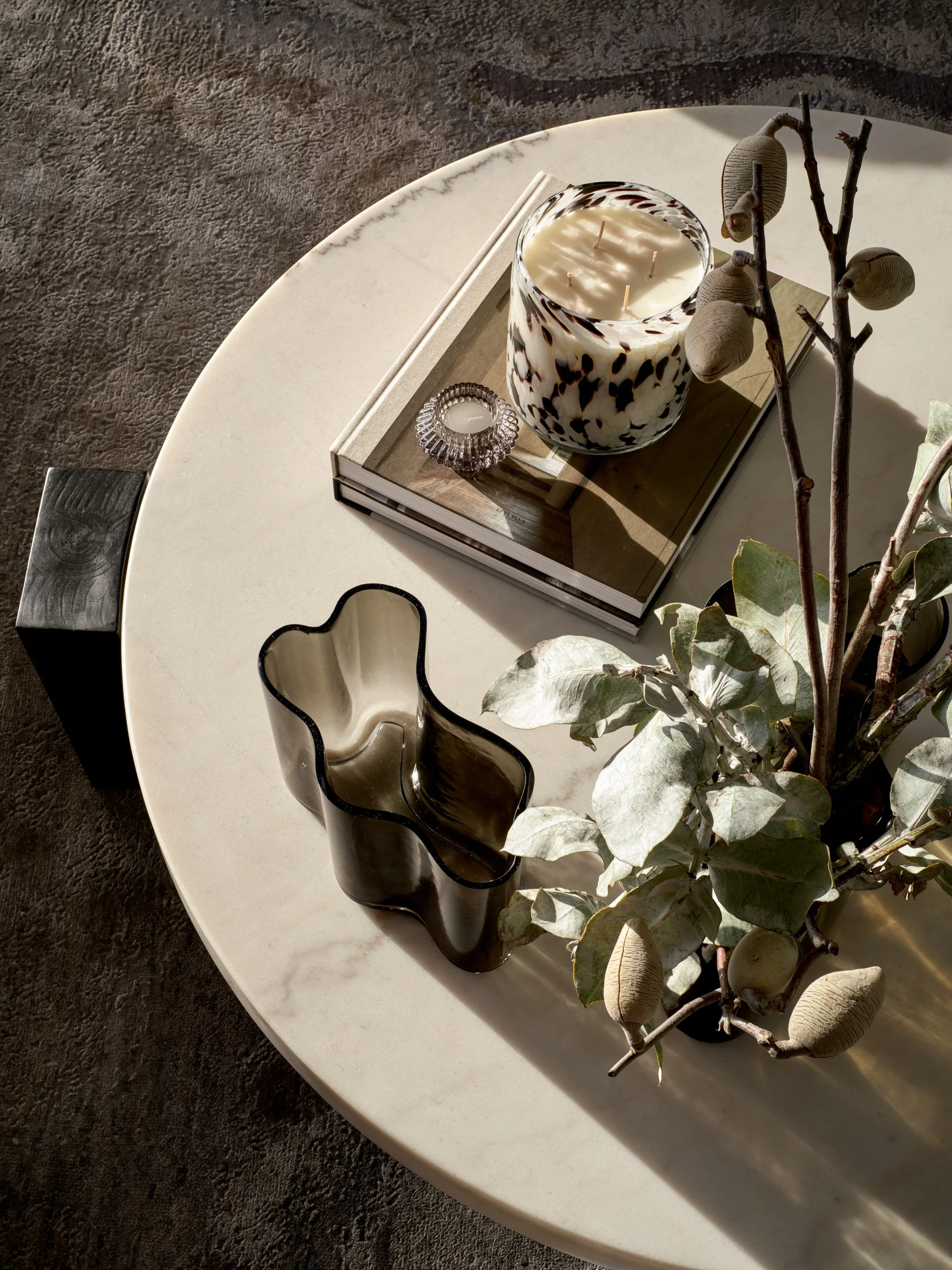 Round marble table with a glass vase, green foliage, candle on a book, and a decorative piece.