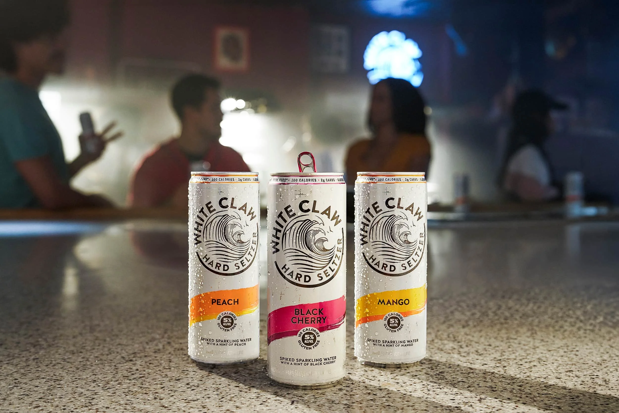 White Claw hard seltzer cans on a bar counter with people in the background, commercial beverage photography