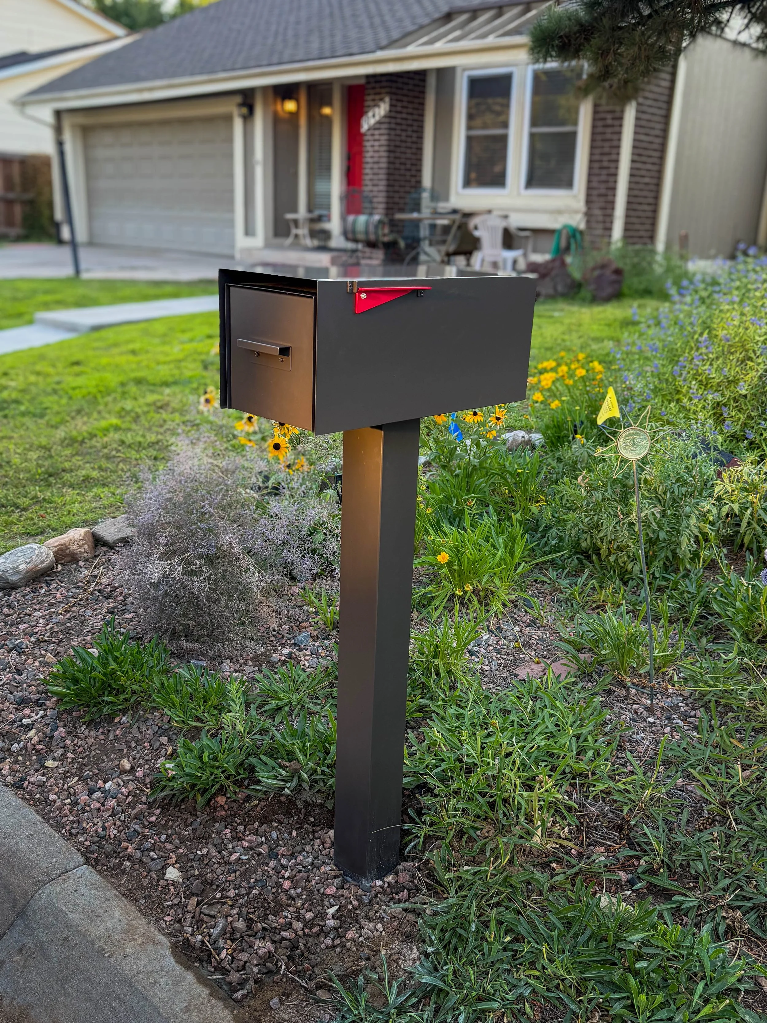 A dark gray mailbox mounted on a dark gray post in a grassy yard with flowers and plants in the background installed by 303 Handyman Services.