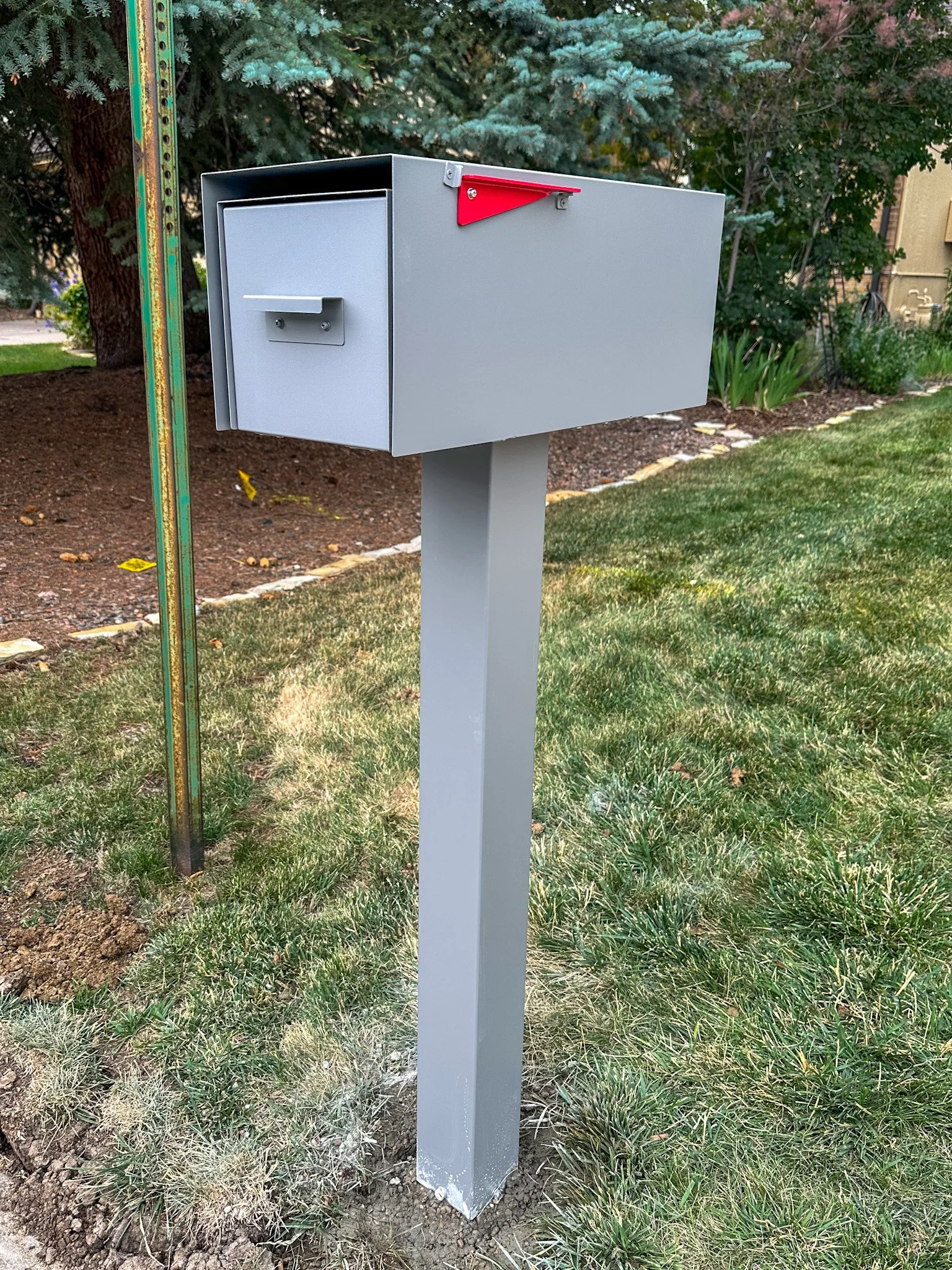 Gray mailbox mounted on a gray pole in a grassy yard with trees and plants in the background installed by 303 Handyman Services.