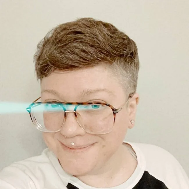 Picture of a white person smiling, with short, brown and grey hair, large-framed tortoiseshell glasses like their dad probably wore in the 1980s, and a medusa piercing.