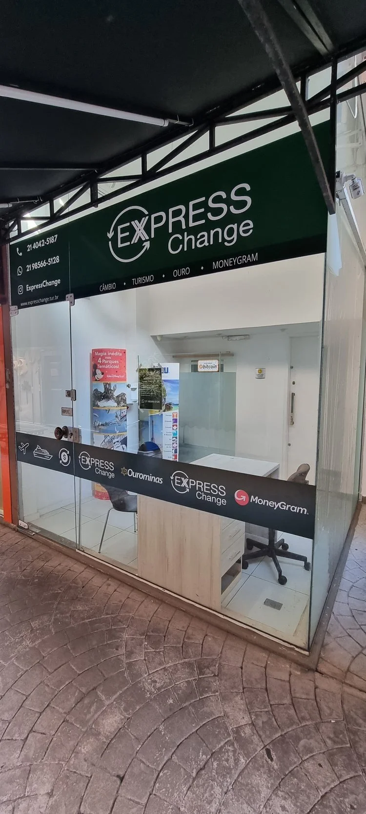 Loja de câmbio Express Change com fachada de vidro, mesas e cadeiras no interior, e cartazes de turismo na parede. Serviços de câmbio, turismo e transferência de dinheiro são oferecidos.