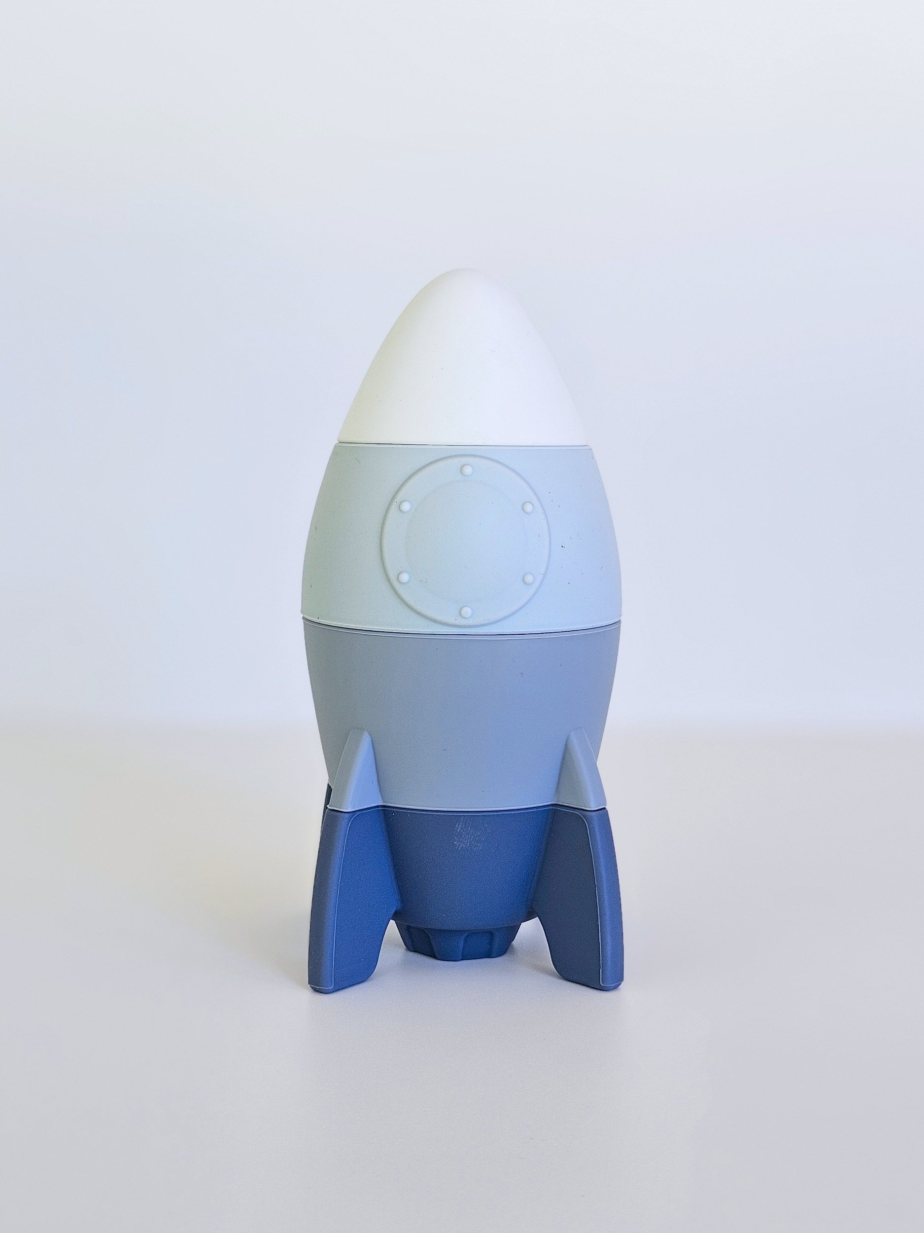 A small toy rocket with three blue fins and a white and light blue body standing on a plain white surface against a white background.