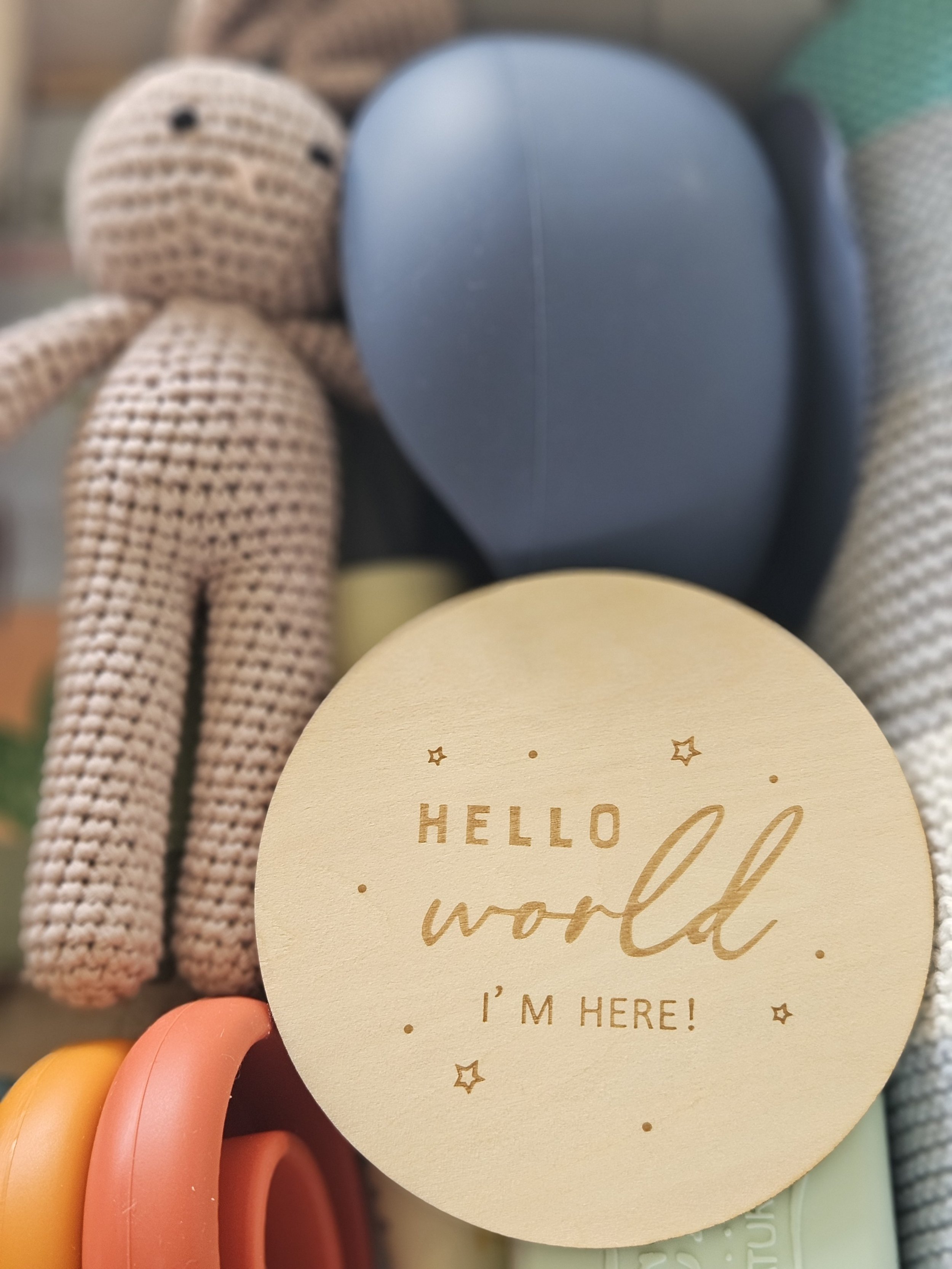 A crocheted teddy bear with black button eyes, surrounded by various objects, including a round yellow card with the words 'Hello world, I'm here!' written on it.