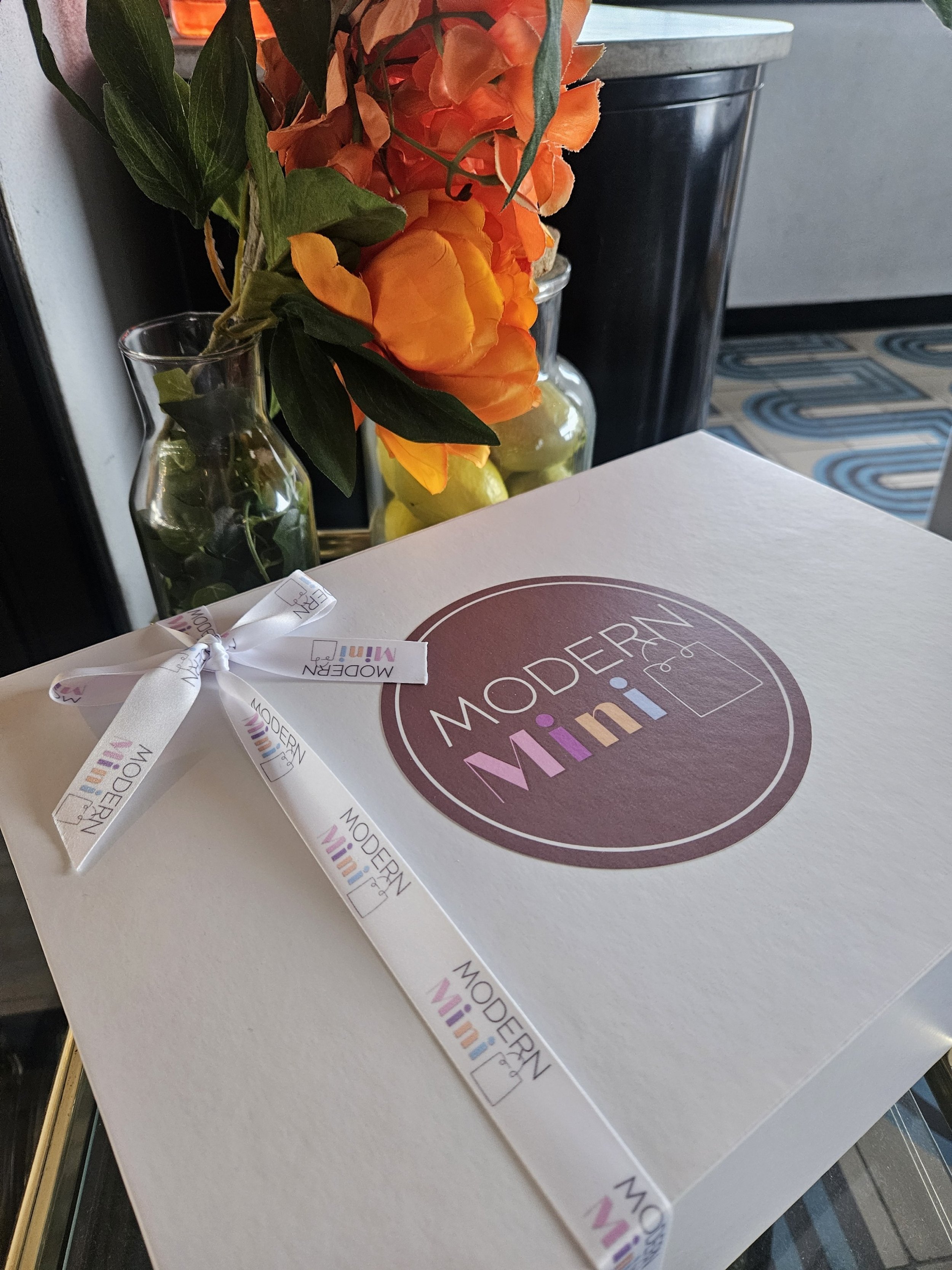 A white box with a circular logo reading 'Modern Mini' and a white ribbon with the same logo, placed on a glass surface. Behind the box, there are two vases with orange flowers and green leaves.