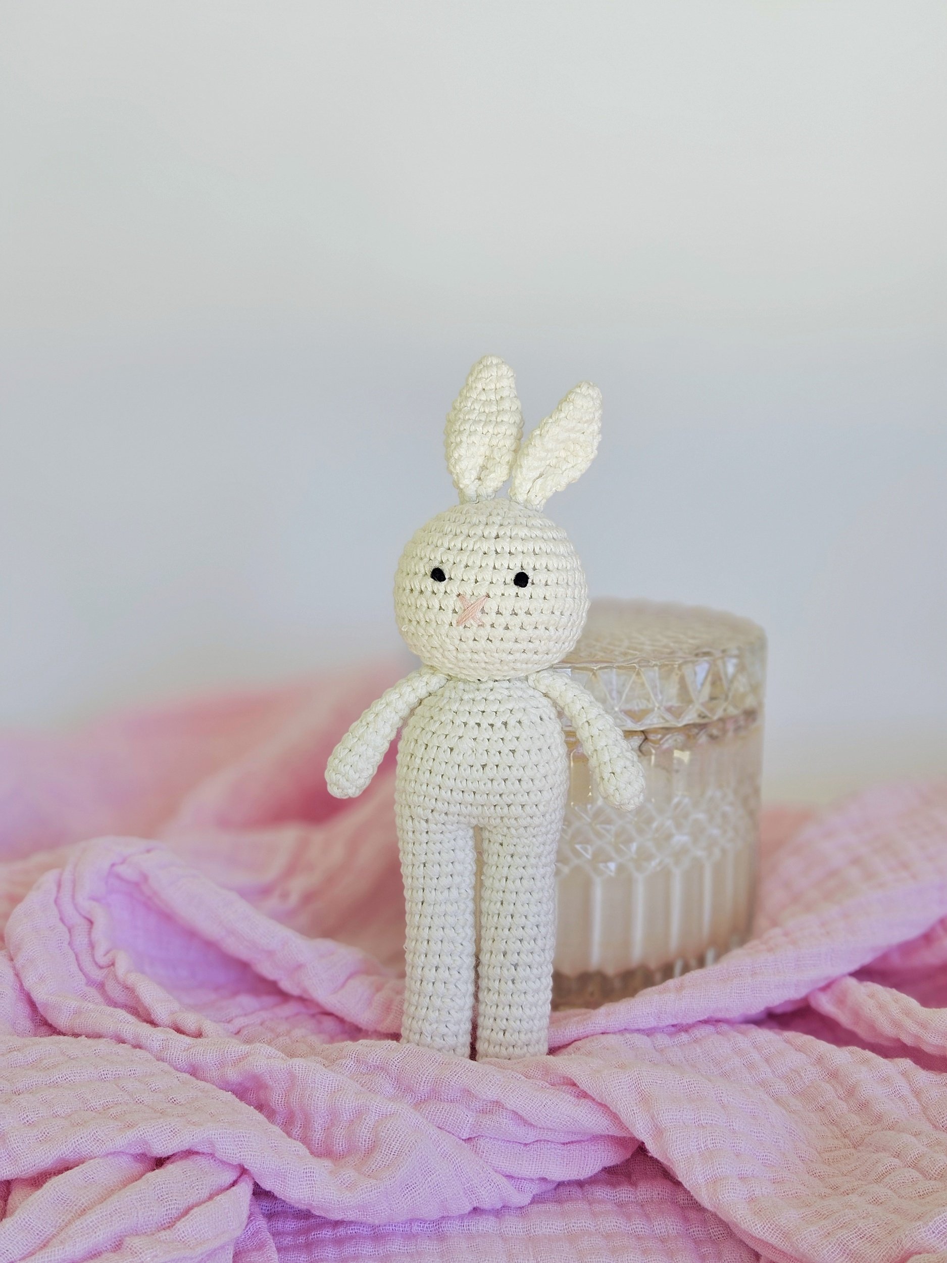 A crocheted white bunny with long ears, black eyes, and a pink nose, standing on a pink cloth. Behind it is a glass container with a textured pattern.