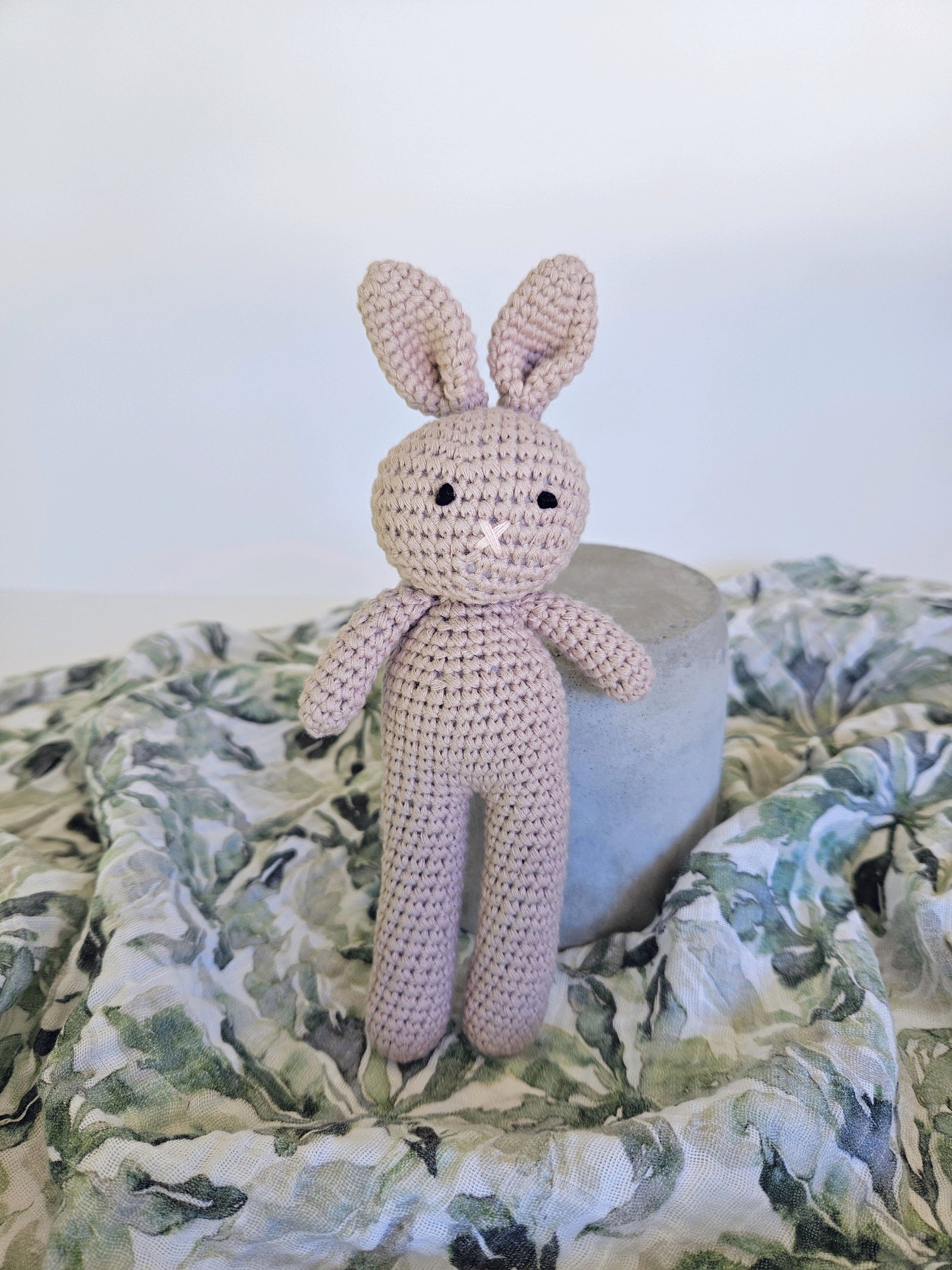 Handcrafted pink crocheted bunny toy with long ears, black eyes, and a small white nose, standing upright on a marble surface draped with a green and white patterned fabric.