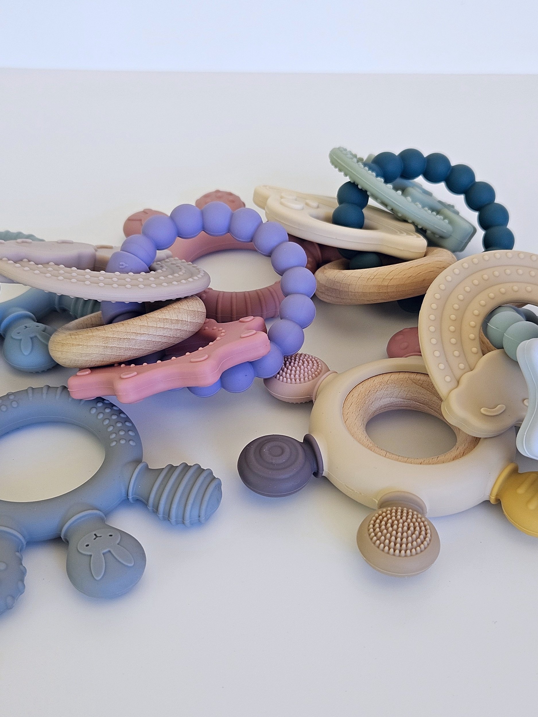 Collection of baby teething toys in pastel colors, including rings, beads, and wooden accessories, scattered on a white surface.
