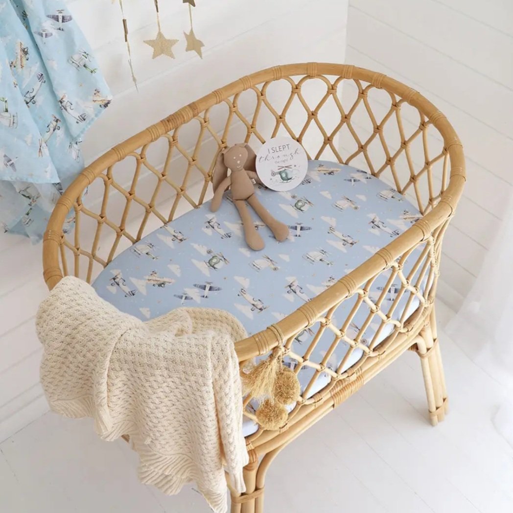 Wicker baby crib with light blue patterned bedding, a beige knitted blanket with pom-poms hanging over the edge, a plush teddy bear, and a round decorative sign on the mattress that says 'I slept'.