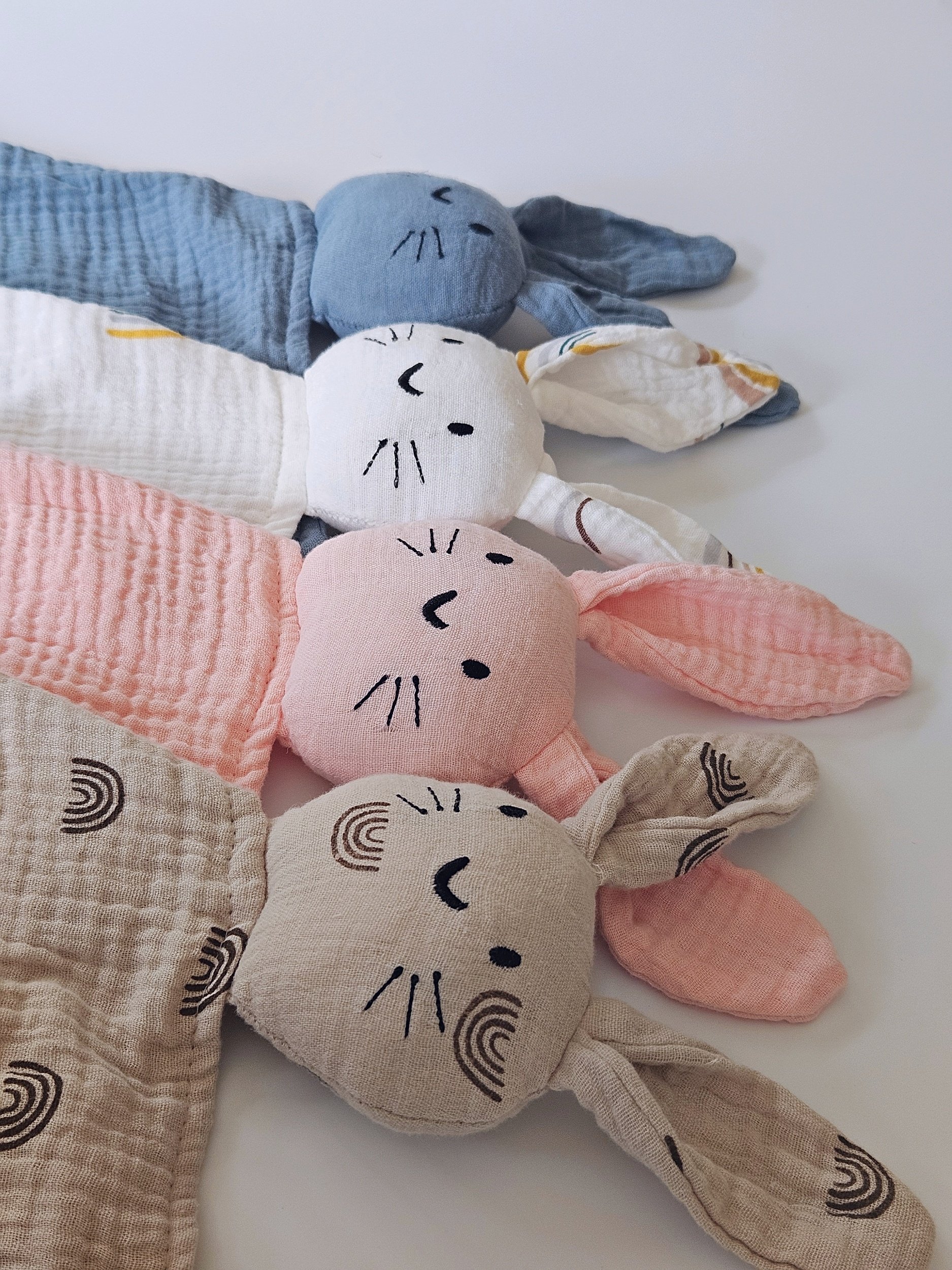 Four plush bunny toys with embroidered faces in blue, white, pink, and beige colors, laid out on a white surface.