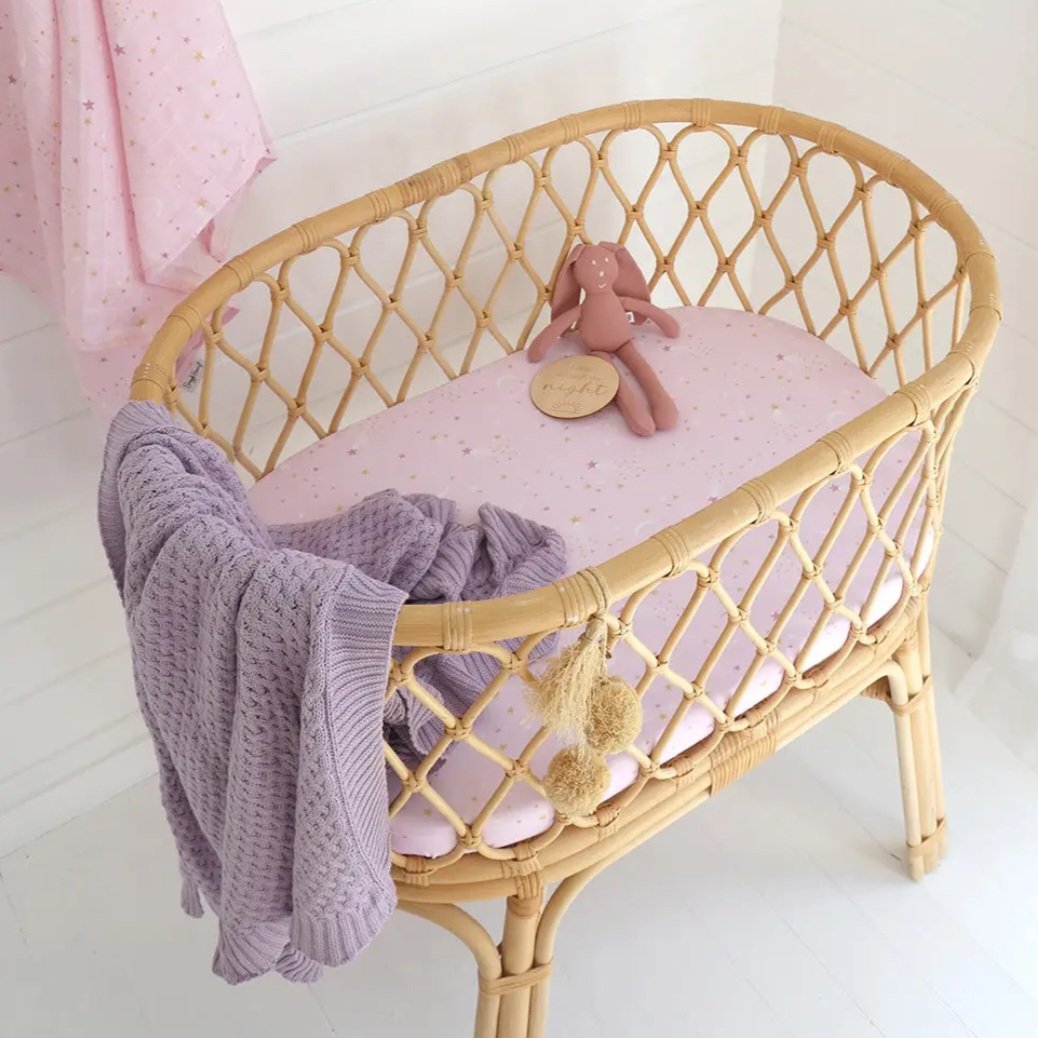 A baby bassinet with pink and purple bedding, a plush bunny toy, a lavender knitted blanket, and a pink curtain with gold stars in the background.