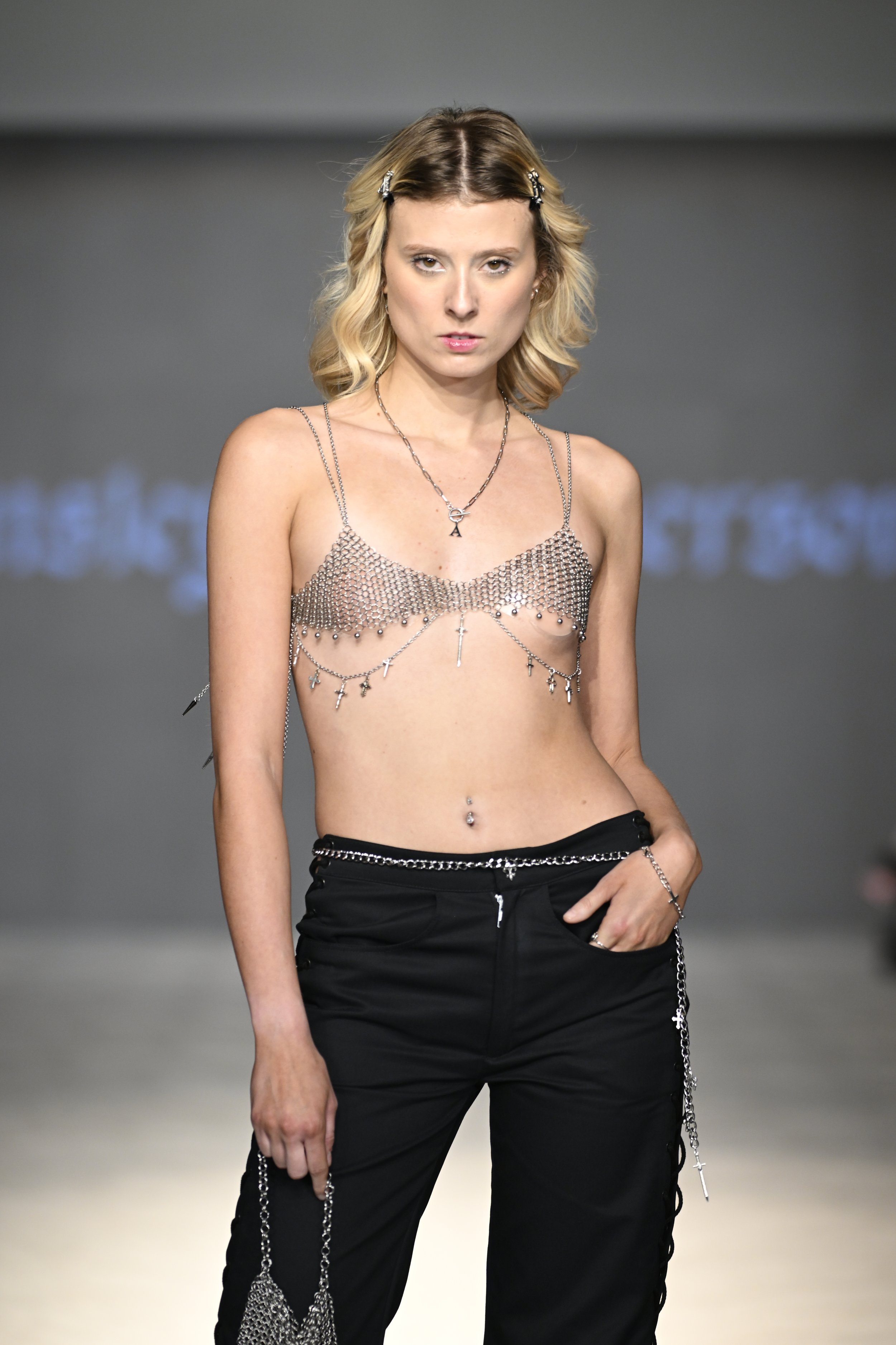 Julia wears a chainmail bra top adorned with chain and charm details, paired with laced low rise jeans in black denim. She carries a matching chainmail mini bag with charms.