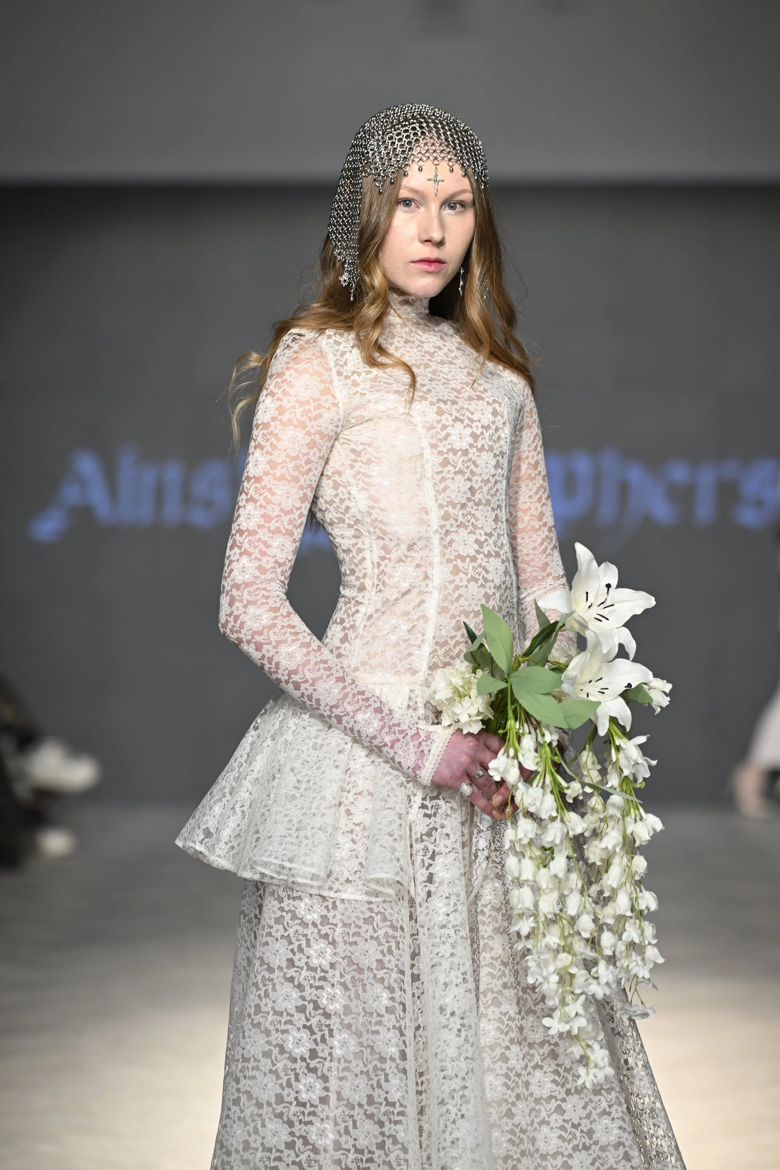 Jasmine wears a corseted drop-waist semi-sheer wedding gown of ivory lace and organza. She wears a handmade chainmail headpiece.