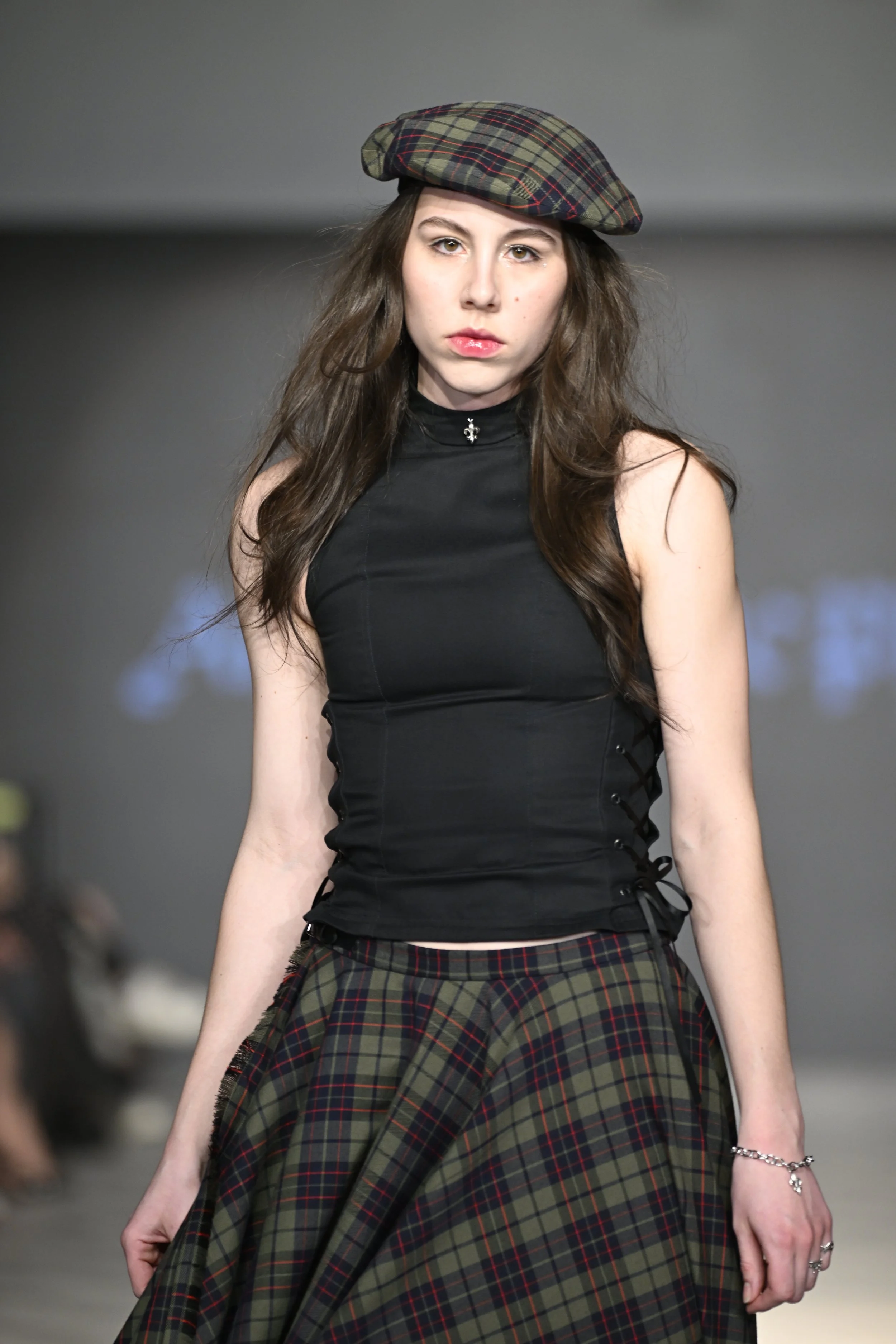 Madeline wears a sleeveless mock neck top with laced details, paired with an asymmetrical handkerchief wrap skirt in an army green tartan with a matching tartan beret.
