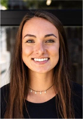Close-up of a young woman with long brown hair, smiling, wearing a black top and a delicate necklace, outdoors with blurred background.