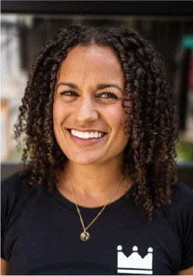 Smiling woman with curly hair wearing a black shirt and a gold necklace, standing outdoors.