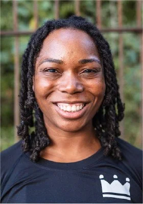 Close-up of a smiling woman with natural curly hair, wearing a black shirt with a white crown logo, outdoors with a blurred green background.